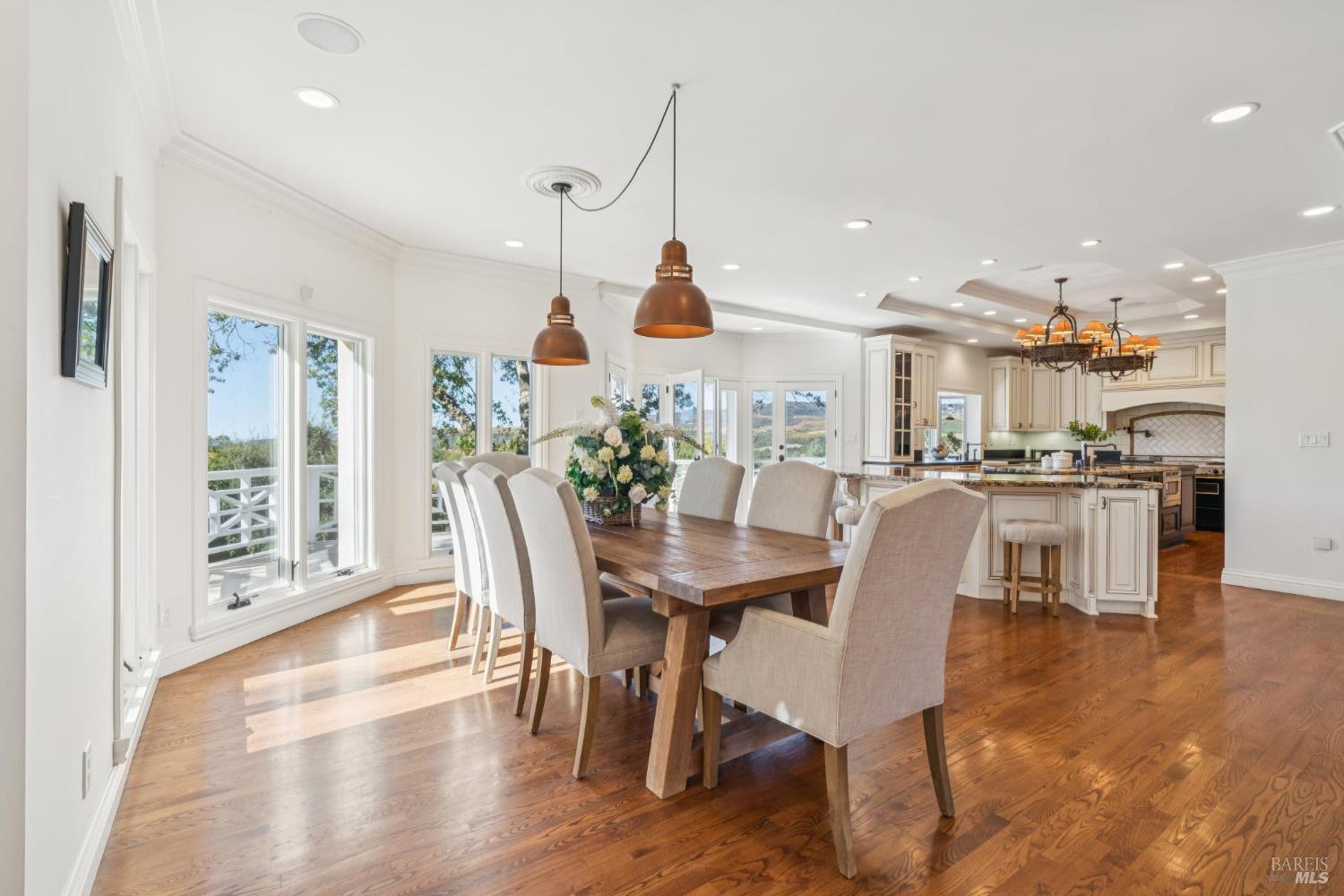Dining room with a long wooden table, upholstered chairs, and an adjoining white kitchen.