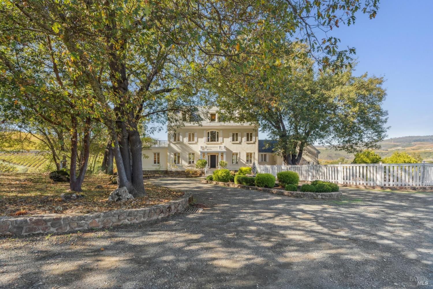 Cream two-story Napa home framed by mature trees, with a white fence and vineyard rows nearby.