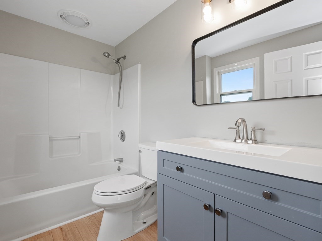 Bathroom with a white tub and shower, blue-gray vanity, and light wood flooring.