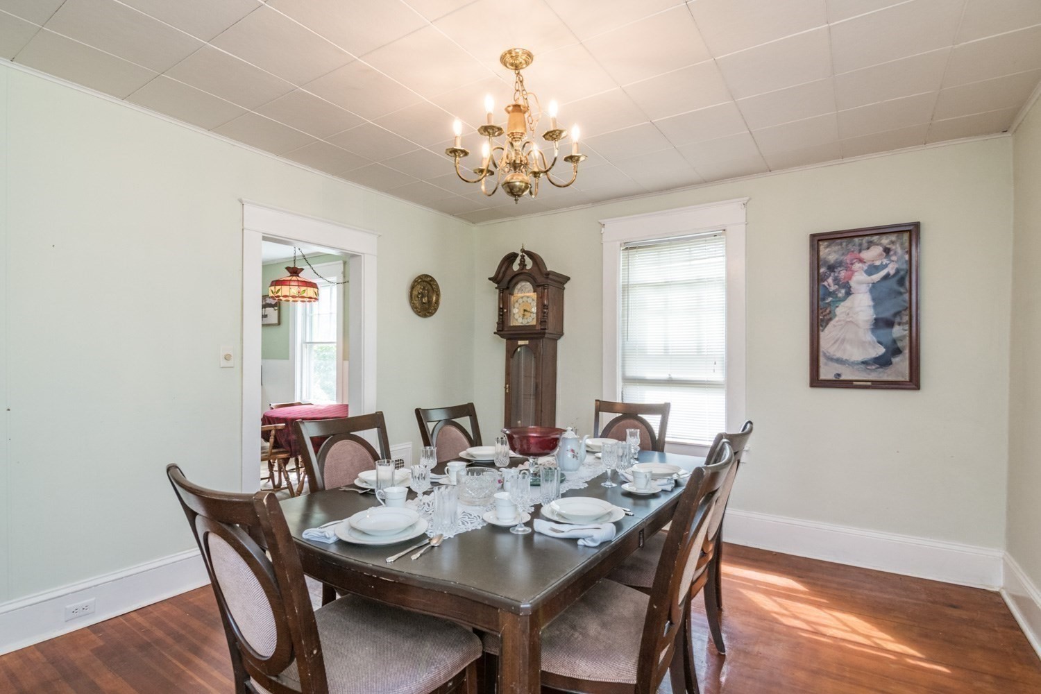 Dining room with a set table, brass chandelier, grandfather clock, and hardwood floors.