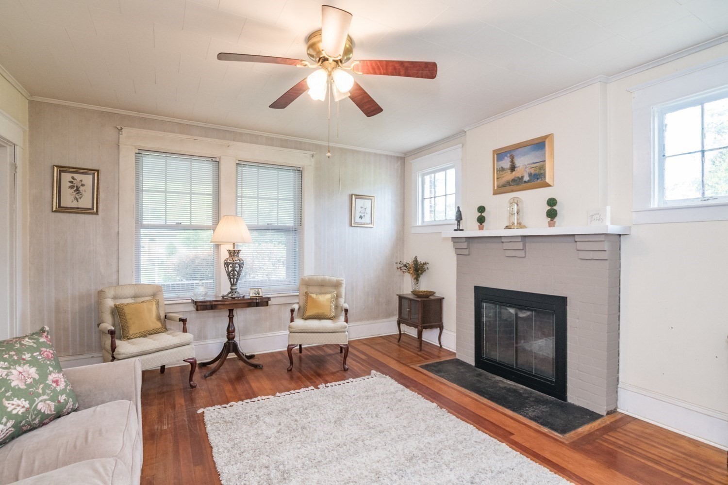 Living room with a painted brick fireplace, hardwood floors, and a ceiling fan with lights.