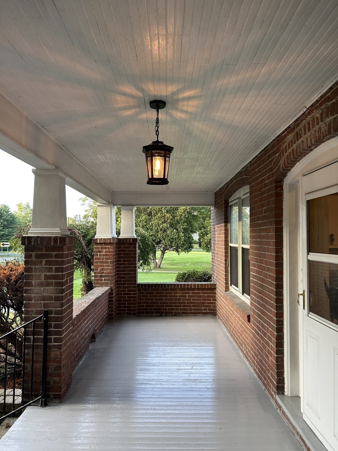 Covered brick porch with white-painted floor, hanging lantern, and a view of the lawn.