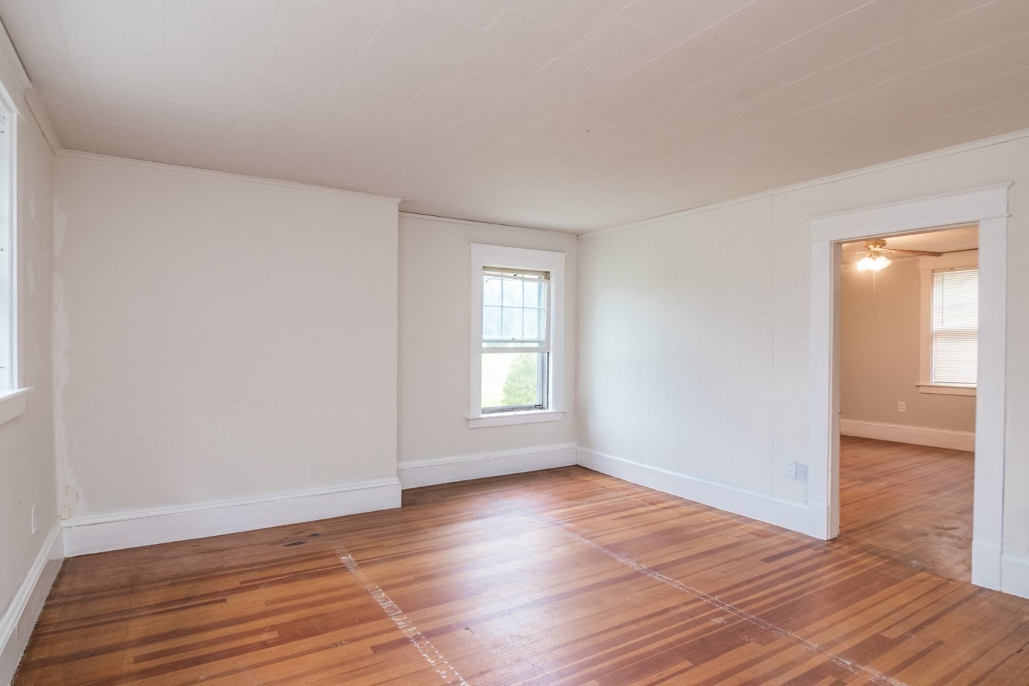 Empty room with striped hardwood floors, white walls, and a doorway to an adjoining room.