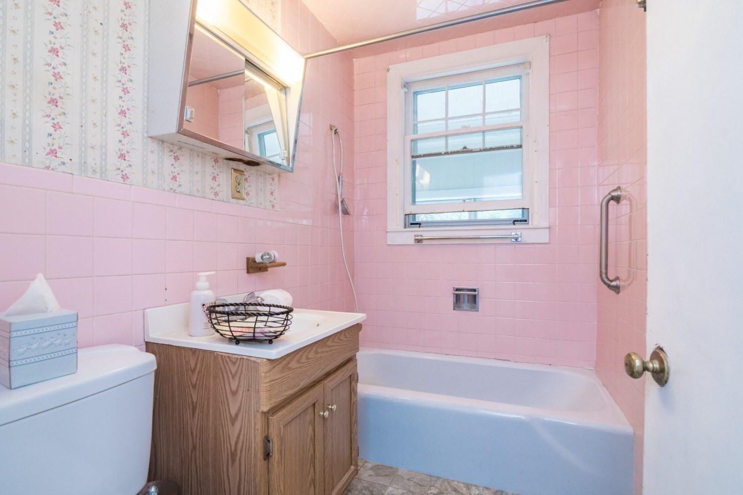 Bathroom with pink tile, a blue bathtub beneath a window, and a wood vanity.