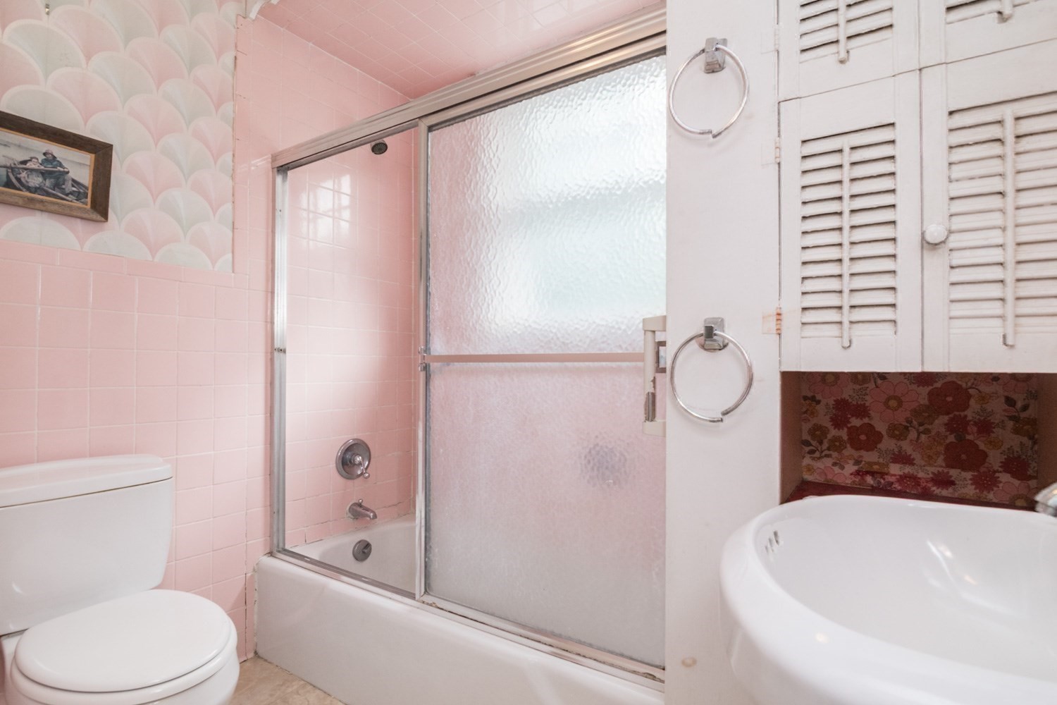 Bathroom with pink tile, frosted-glass tub enclosure, white louvered cabinets, and floral wallpaper.