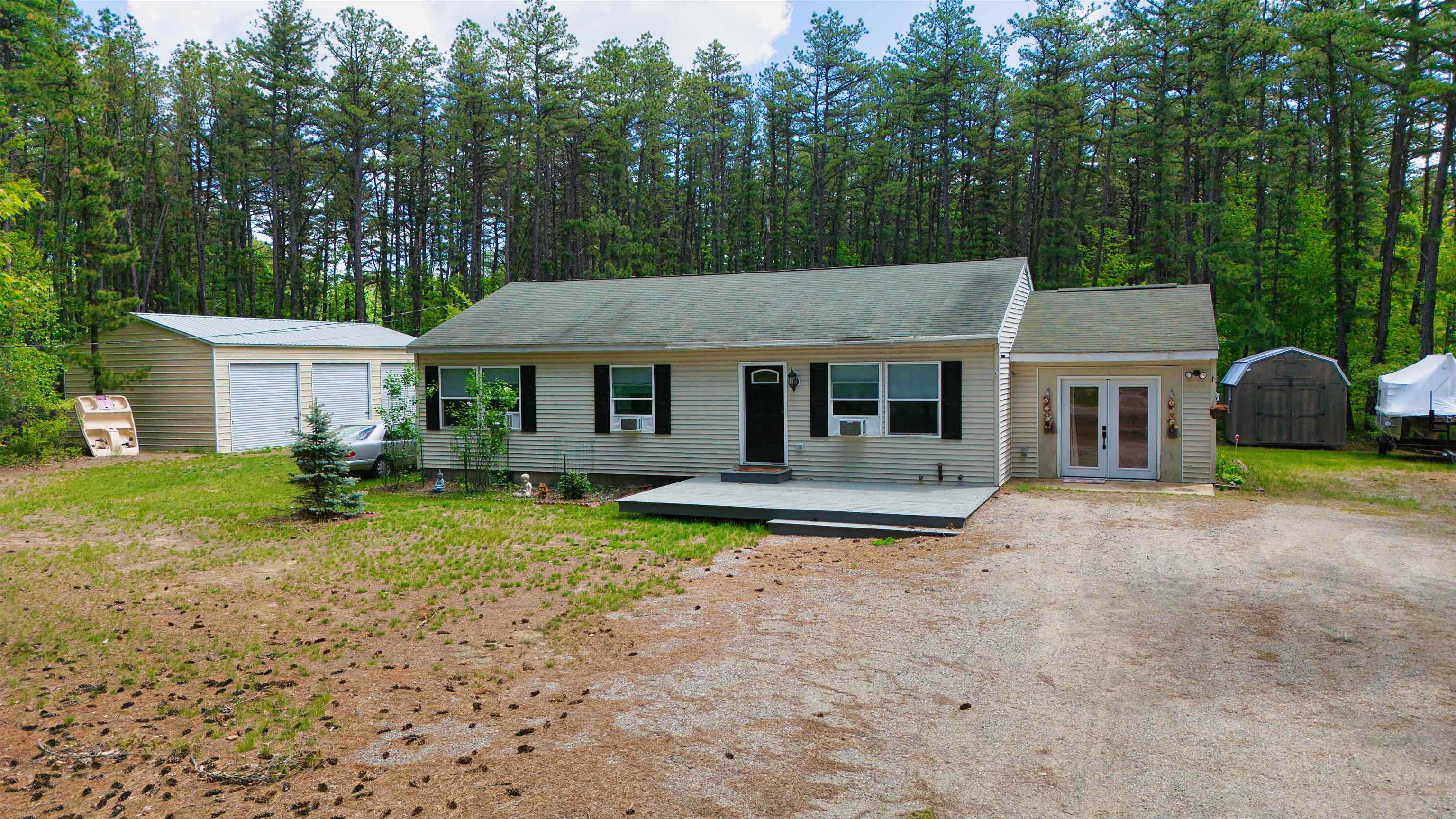 Single-story beige home with a front deck and gravel driveway, surrounded by tall trees.