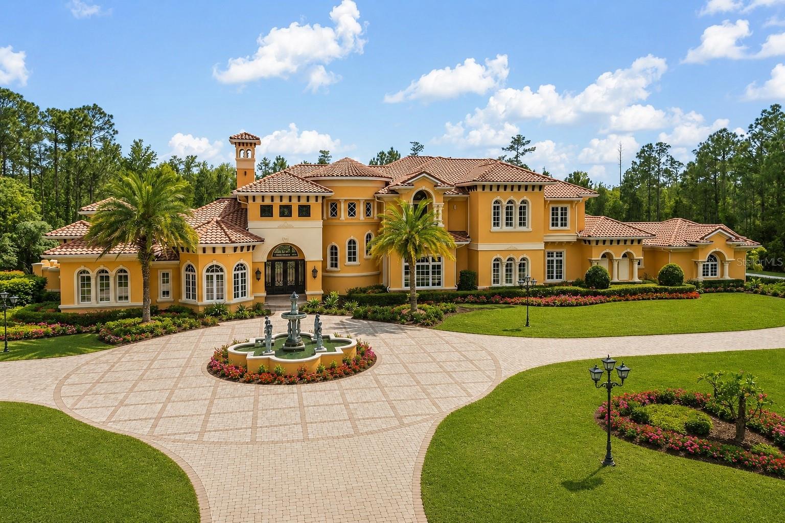 Large golden stucco home with terracotta roofs, a circular drive, fountain, and landscaped lawn.