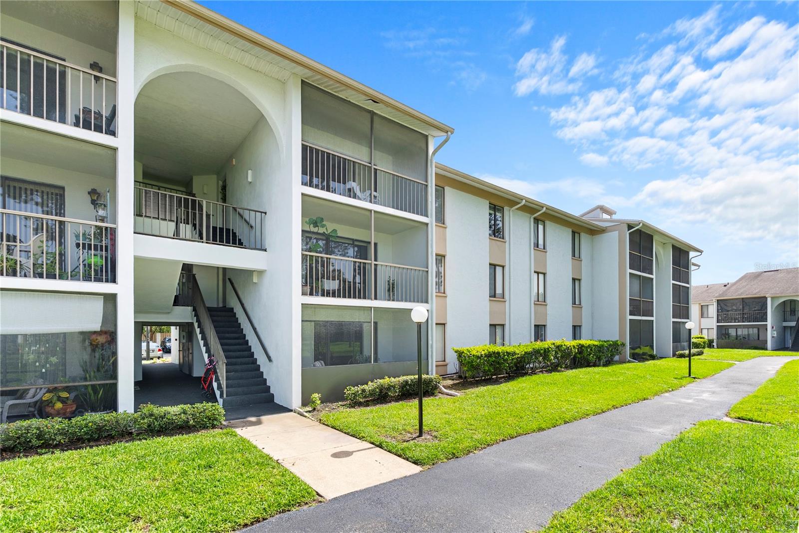 Multi-story condominium building with screened balconies, exterior stairs, and a paved walkway across the lawn.