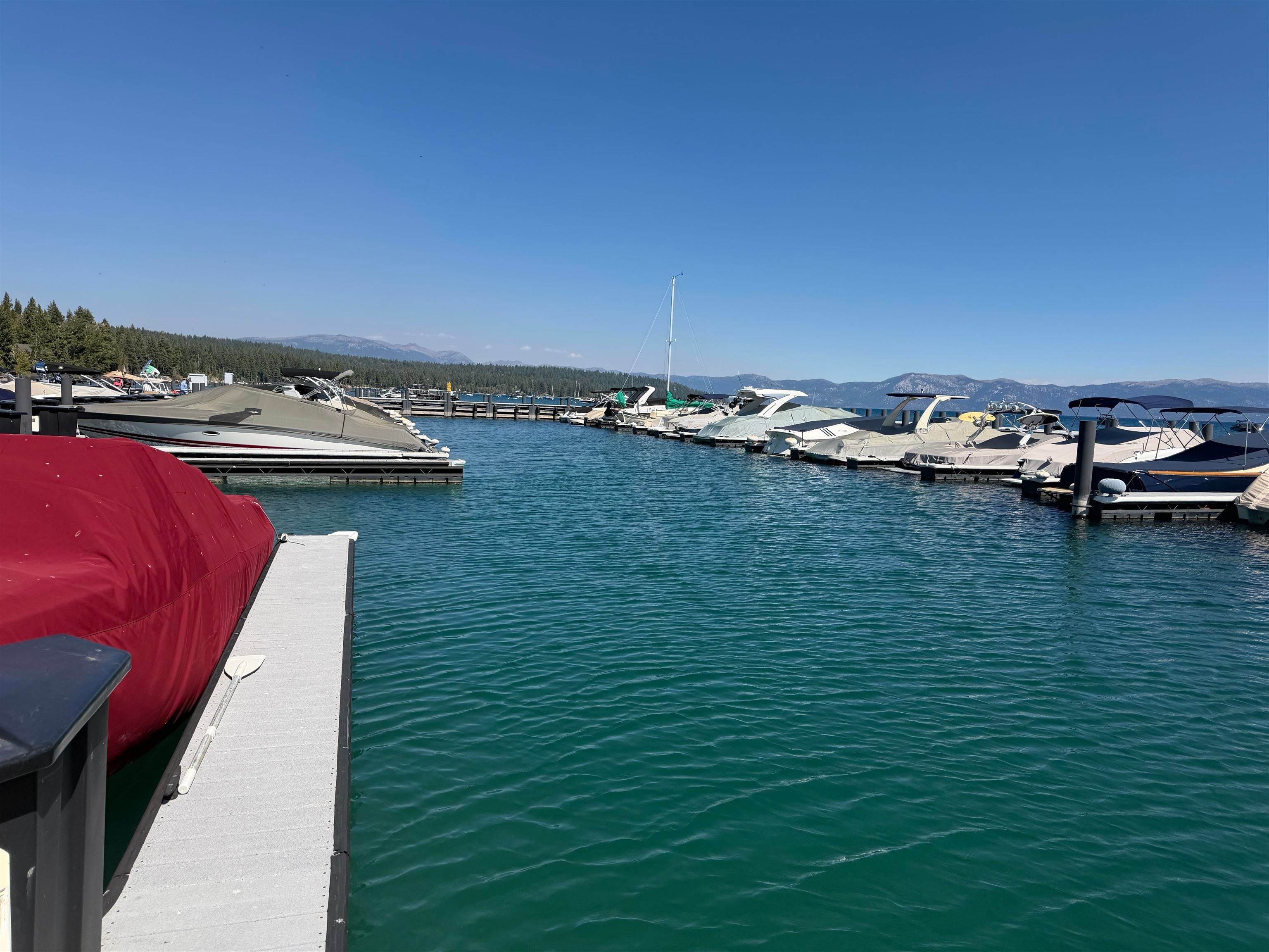 Marina with boats along floating docks on turquoise lake water, backed by forested shores and distant mountains.