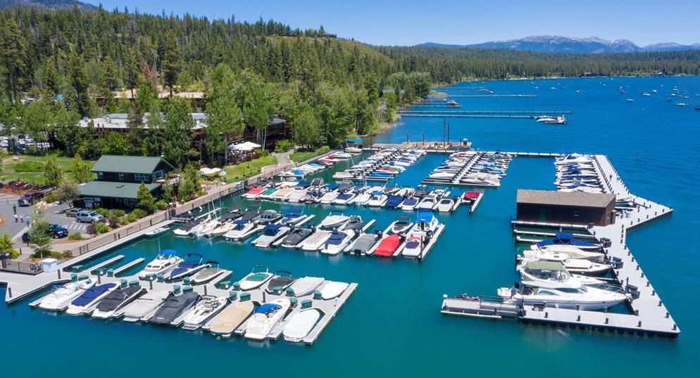 A lakeside marina with rows of boats, forested shoreline, and distant mountains.