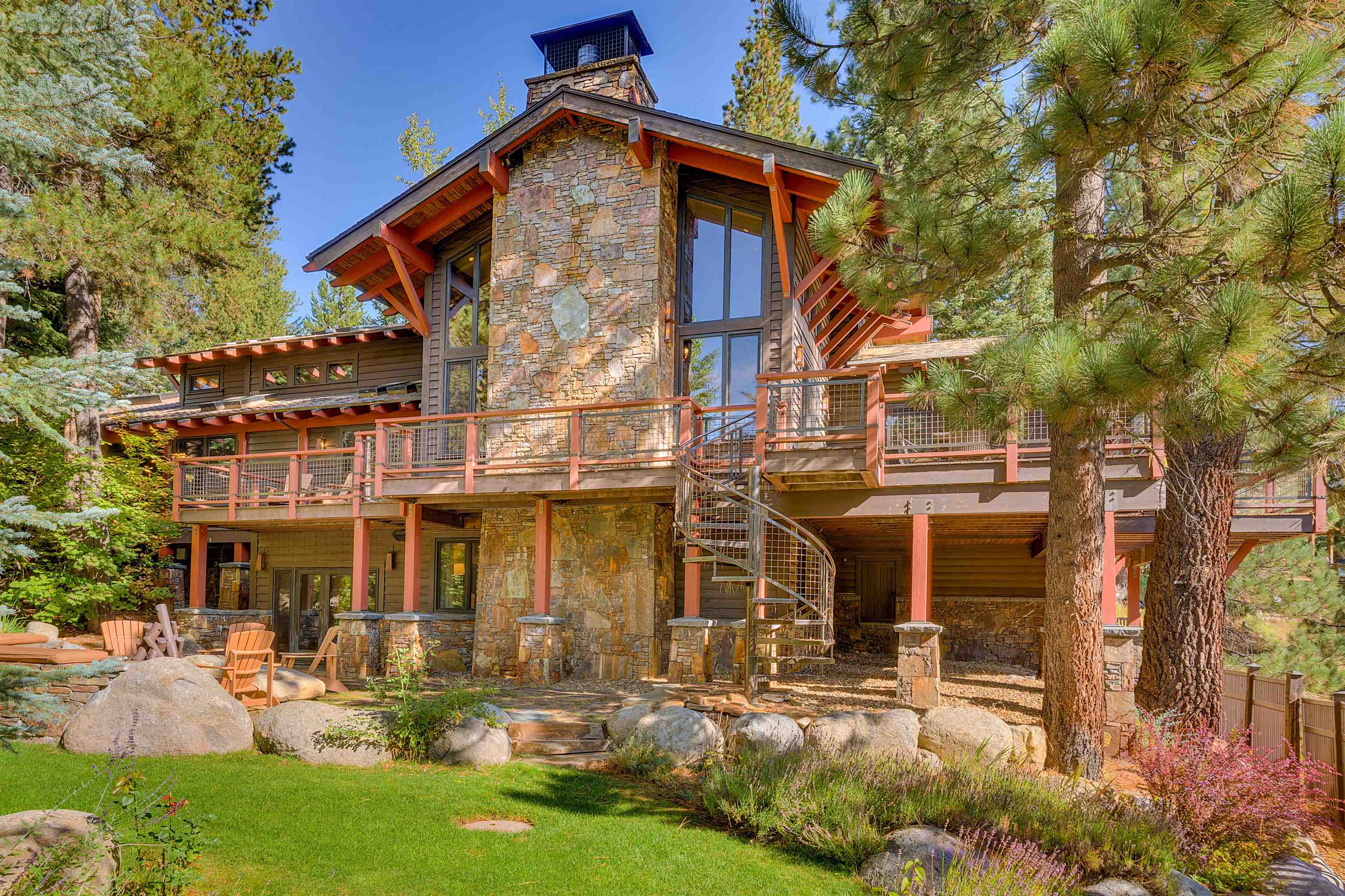 Mountain home with stone chimney tower, expansive decks, and spiral staircase among pine trees.