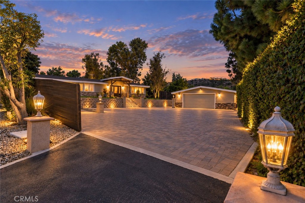 Modern home and detached garage beyond a wide paved driveway, framed by illuminated landscaping at dusk.