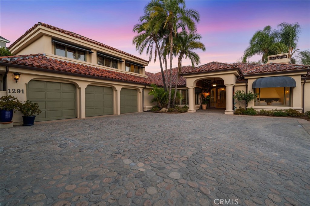 Mediterranean-style home with three garage doors, palm trees, and a wide cobblestone driveway at dusk.