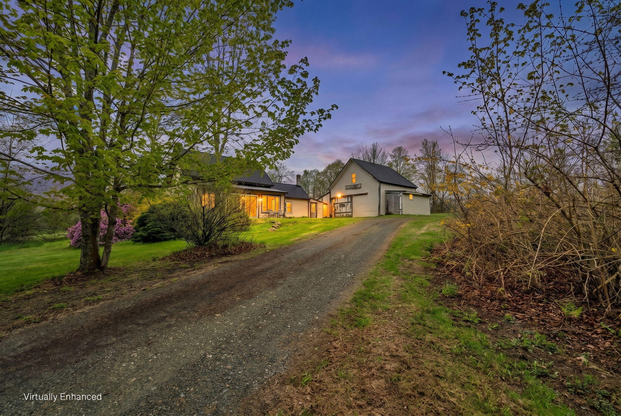 Rural home and outbuilding at dusk, viewed from a gravel driveway amid budding trees and flowering shrubs.