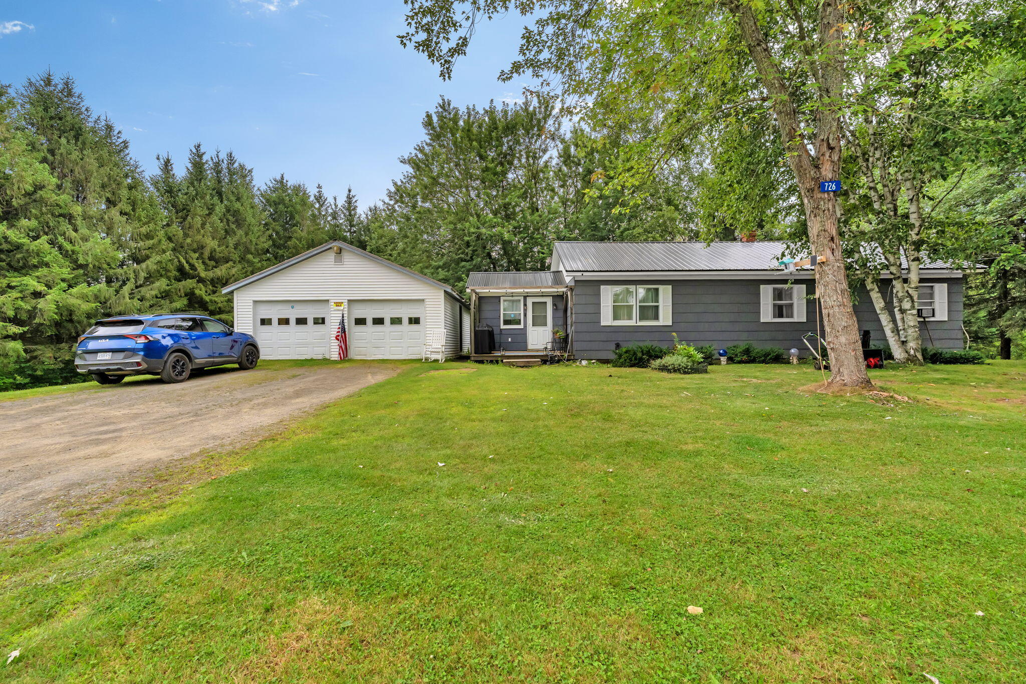 Gray single-story house with a white two-car garage, gravel driveway, and large leafy trees.