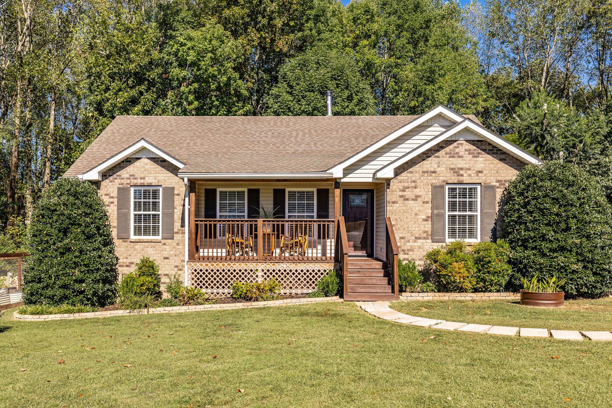 Brick house with a covered wooden porch, landscaped lawn, and mature trees behind it.
