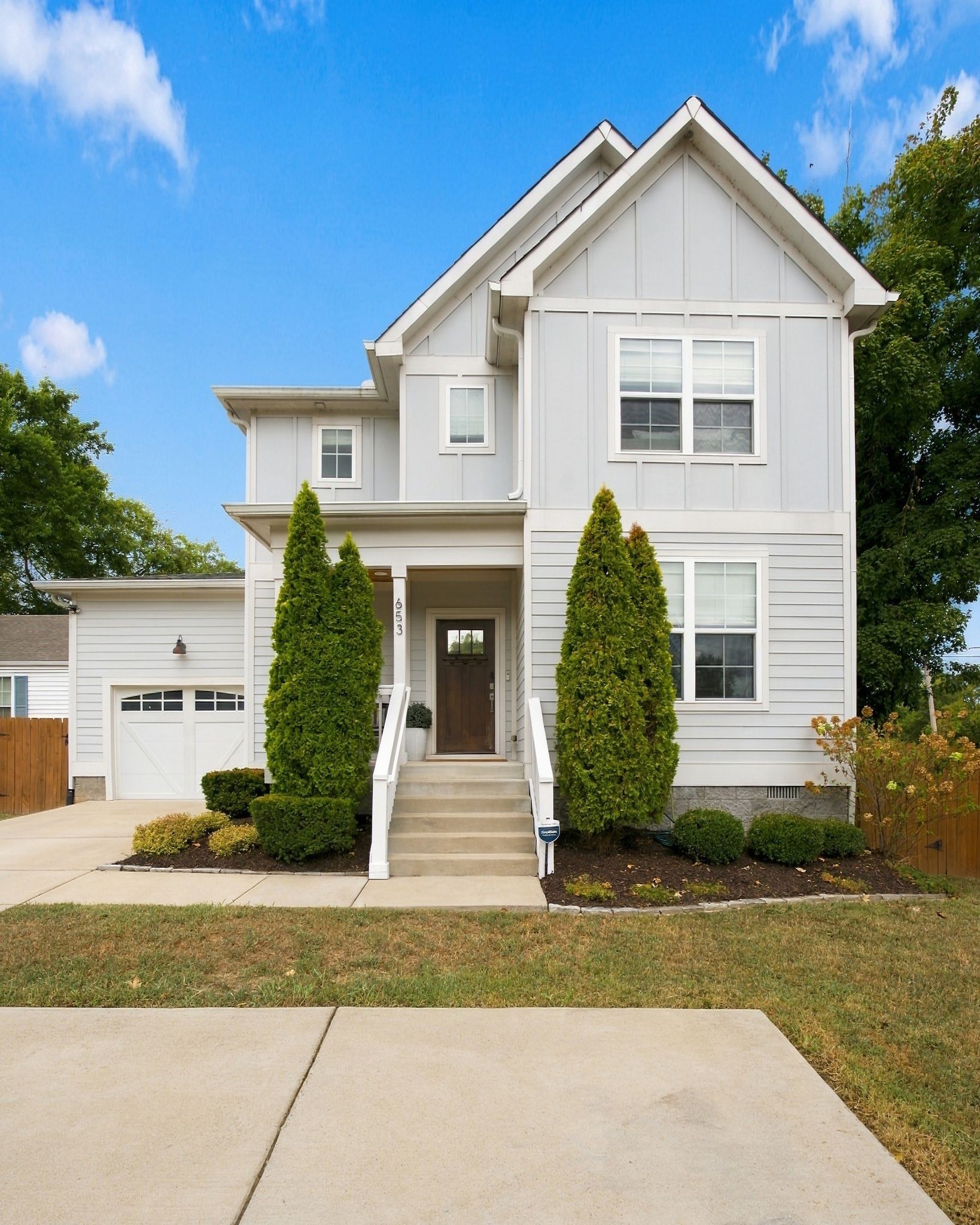 Two-story gray house with a steep gabled roof, attached garage, and evergreen shrubs flanking the front steps.