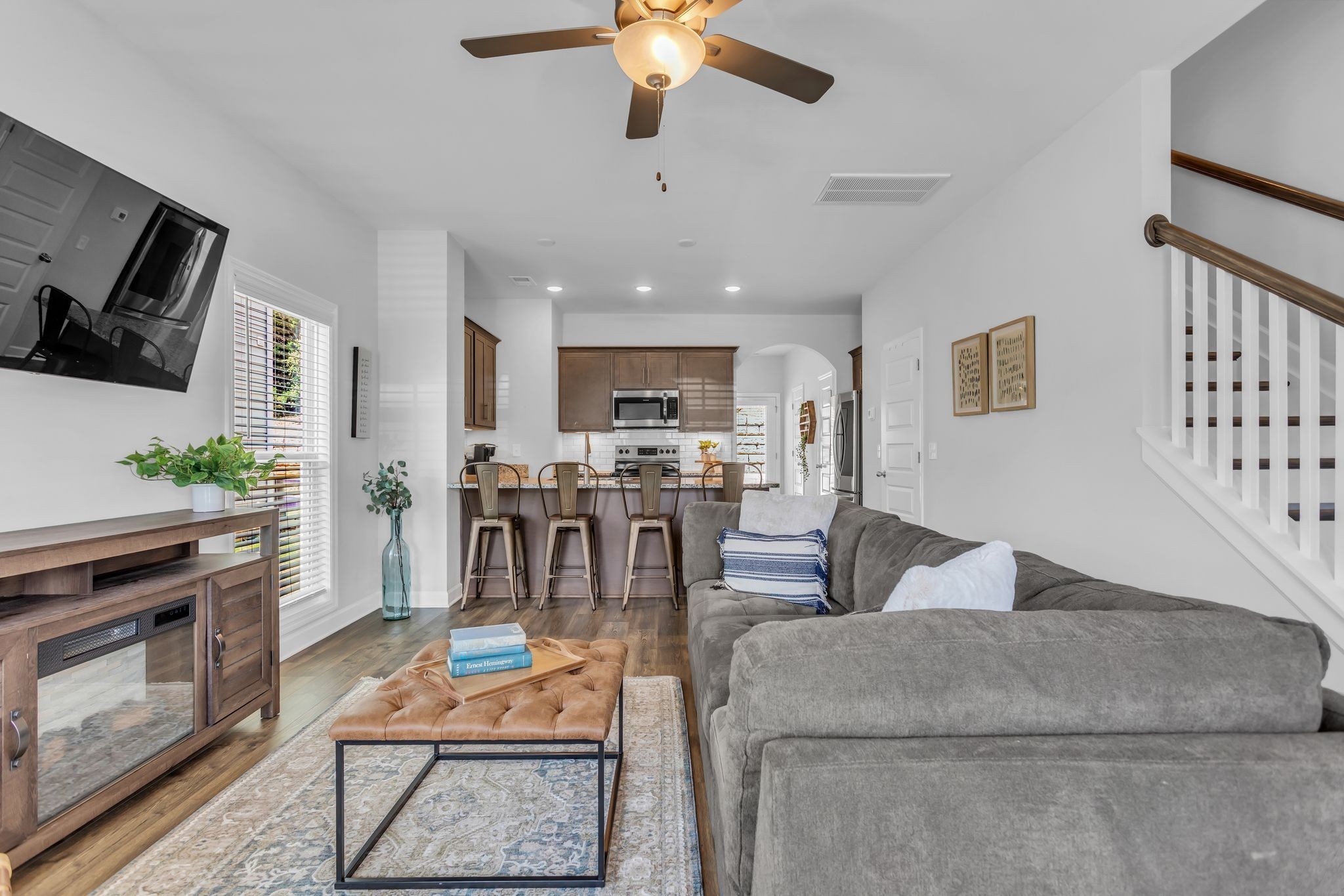 Living room with a gray sectional, leather ottoman, open kitchen, and staircase.