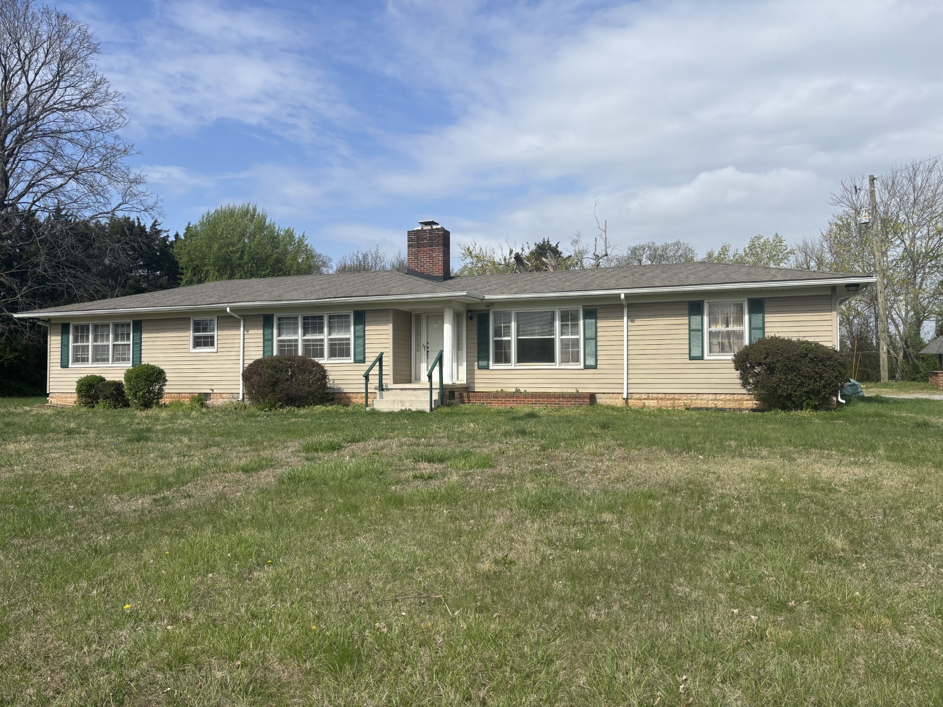 Single-story beige ranch house with green shutters, a brick chimney, and a broad front lawn.