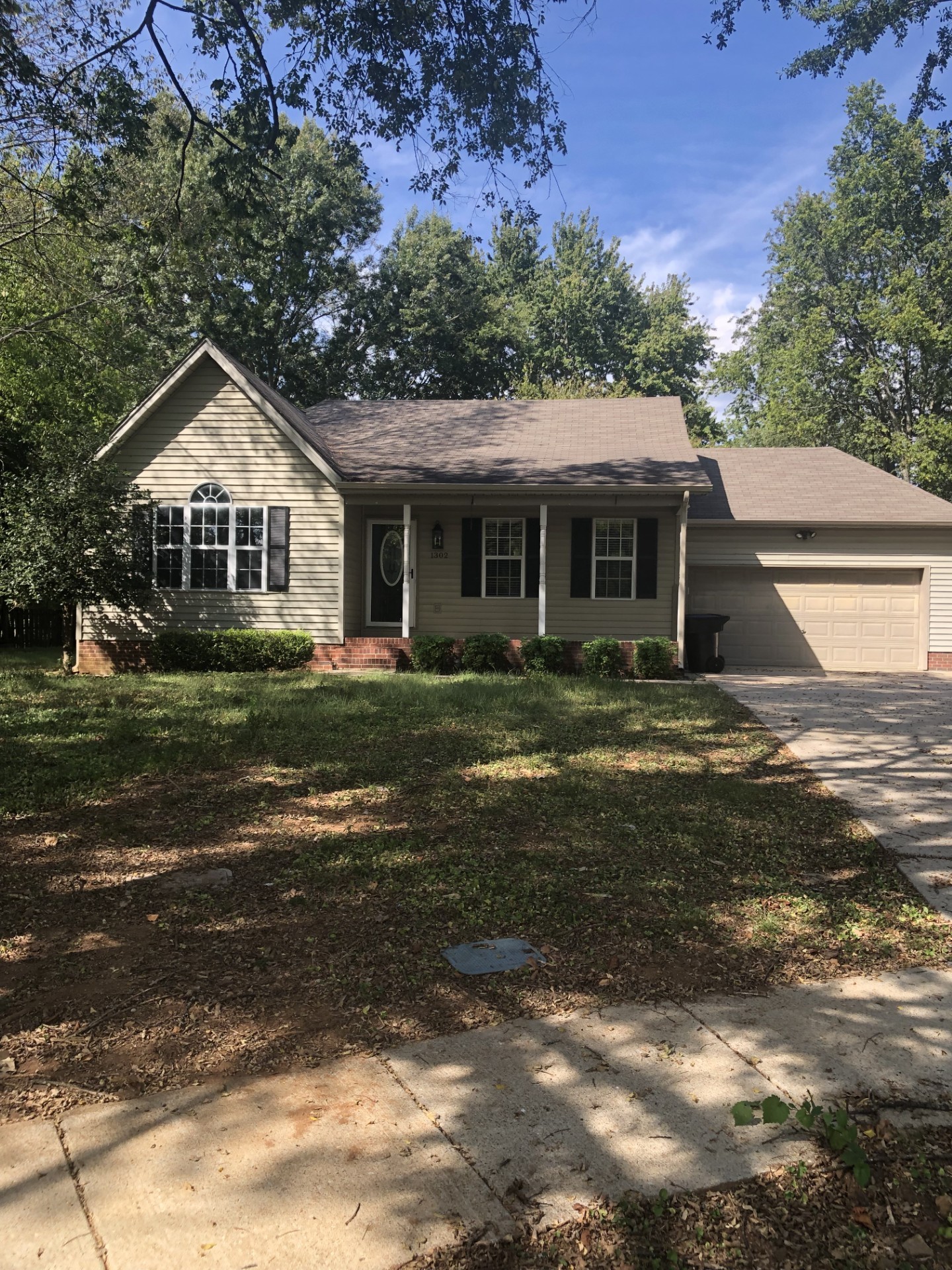 Single-story beige house with an arched front window, covered porch, and attached garage beneath mature trees.