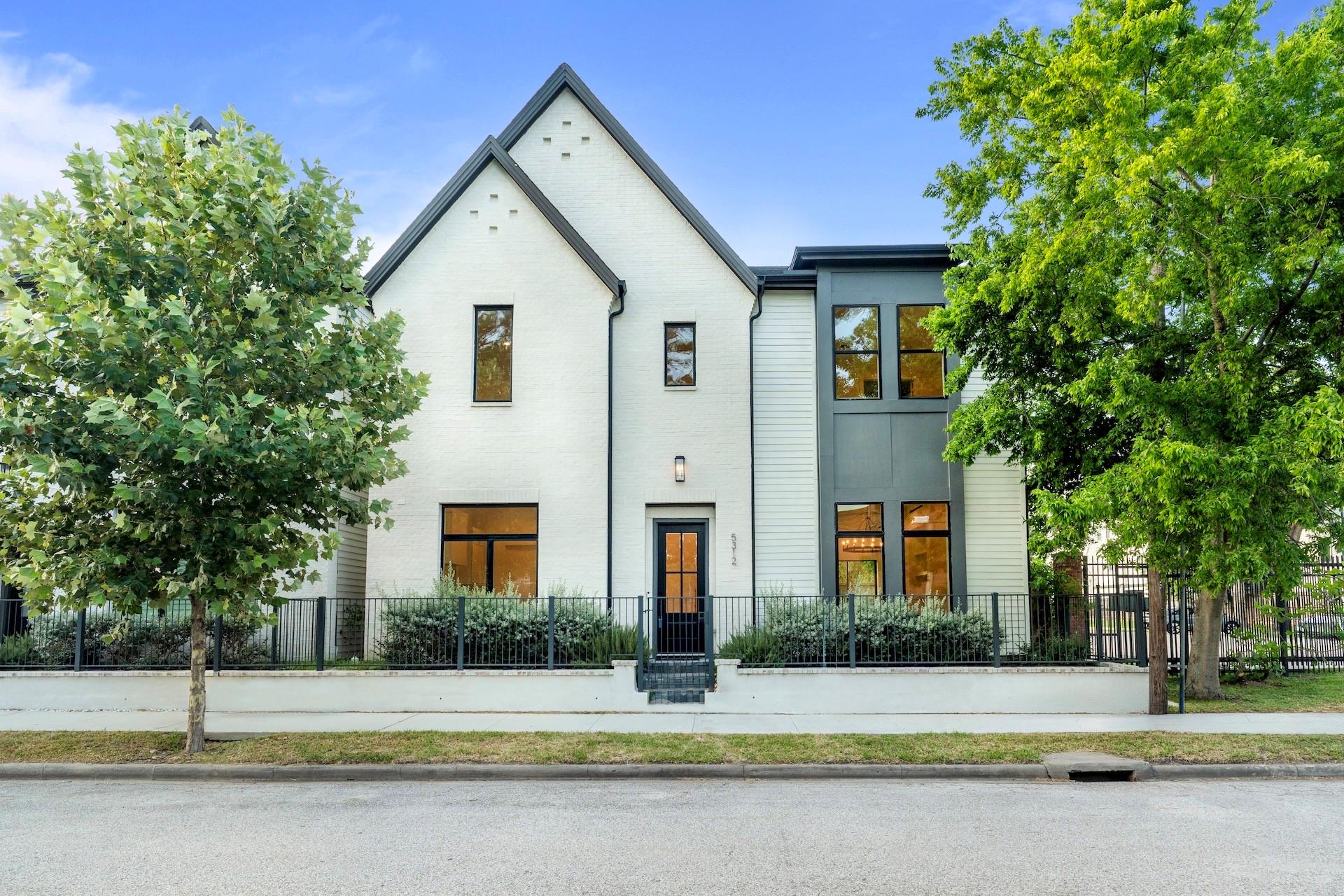 Modern two-story white brick home with steep gables, dark trim, and mature trees along the front fence.