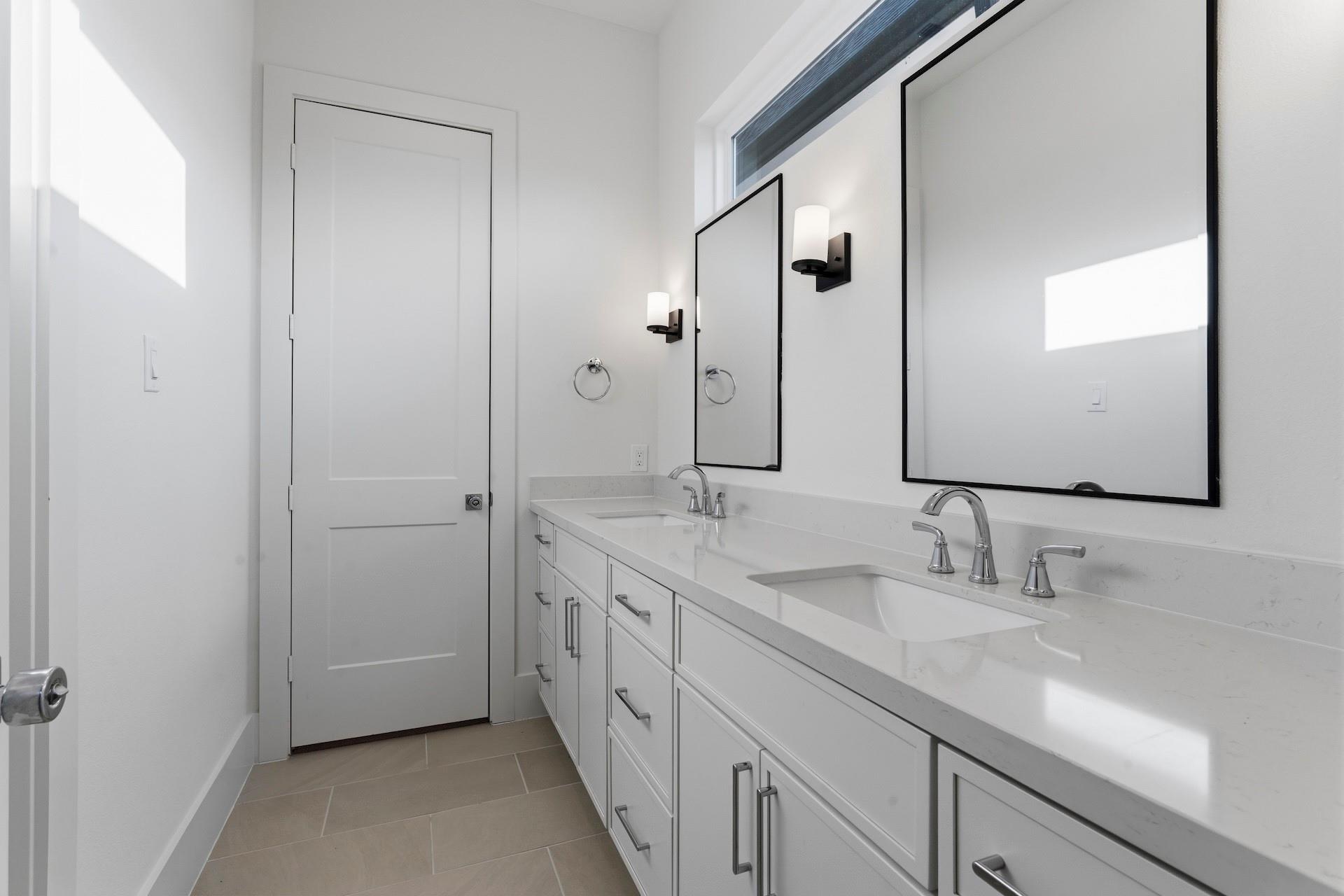 Bright bathroom with a white double-sink vanity, large black-framed mirrors, and beige tile flooring.