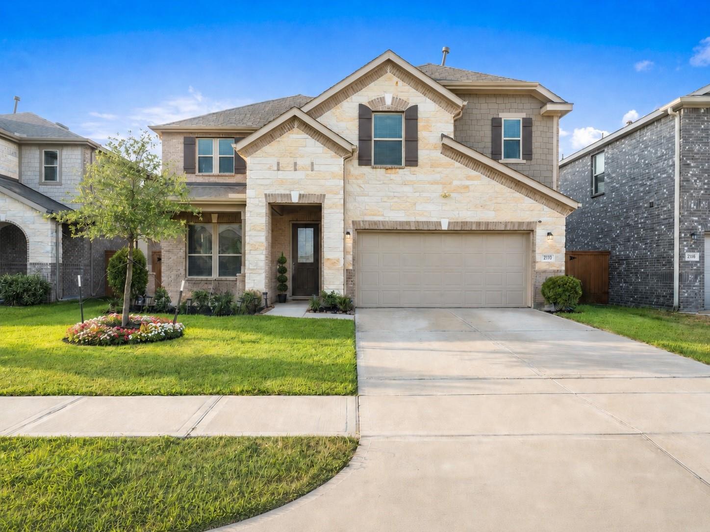 Two-story stone house with a two-car garage, landscaped lawn, and curved driveway.