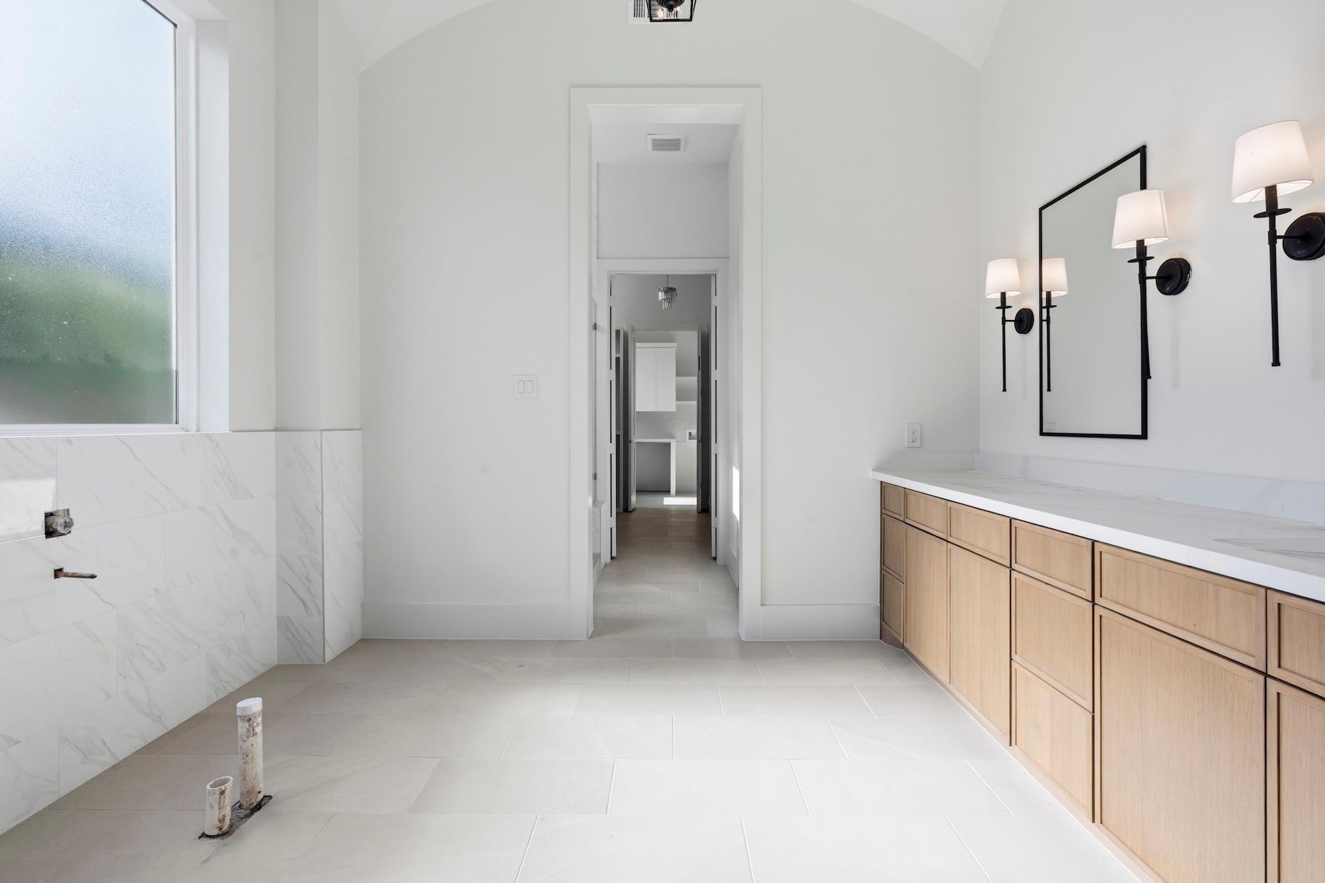 Bright bathroom with a light-wood vanity, marble-look tile, black sconces, and exposed plumbing pipes.