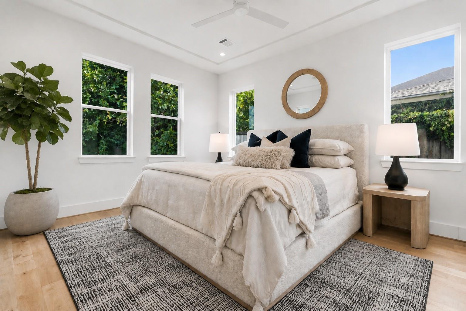 Bright staged bedroom with a cream bed, round wood-framed mirror, patterned rugs, and leafy views through the windows.