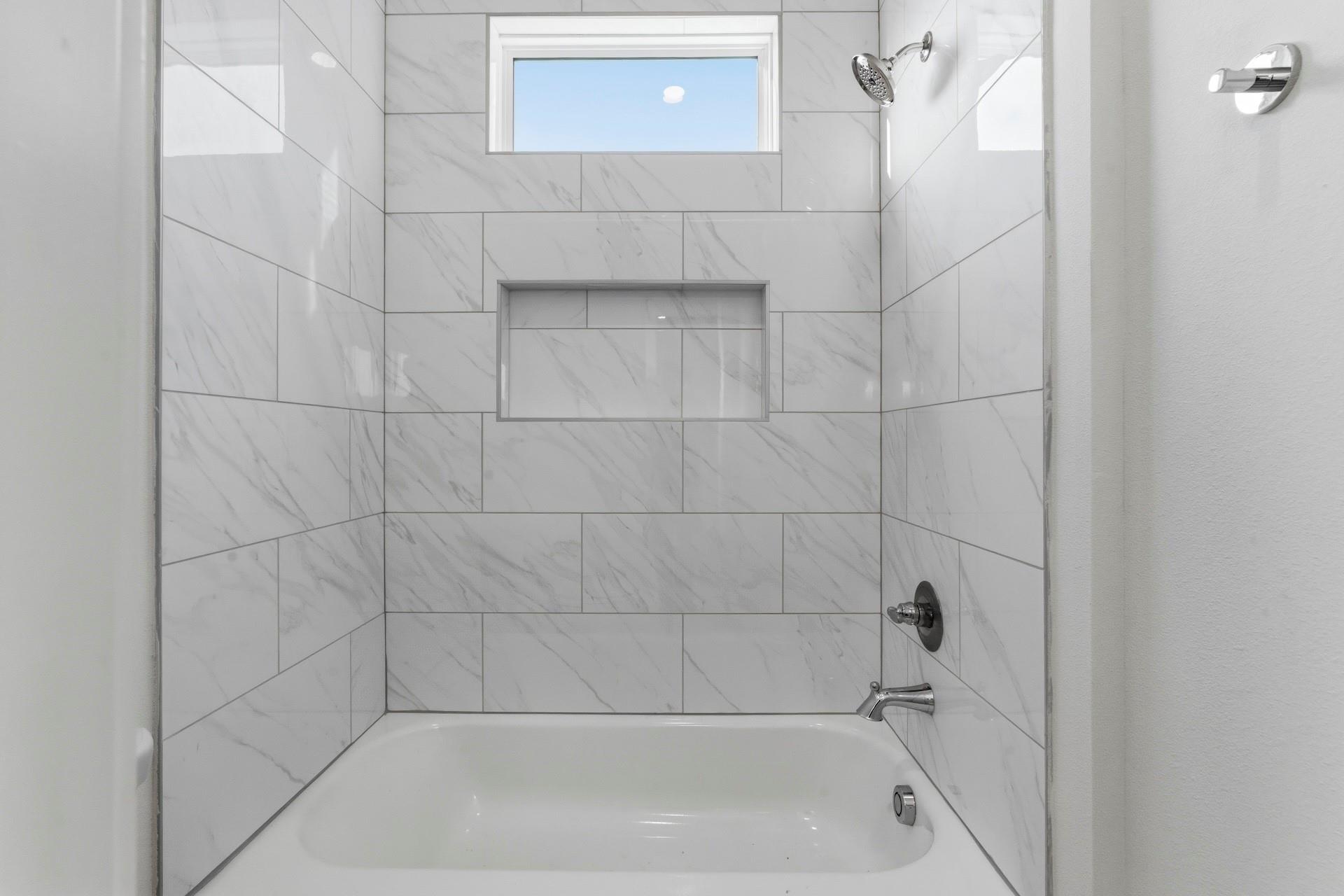 Bathtub in a marble-look tiled alcove with a recessed niche and high window.