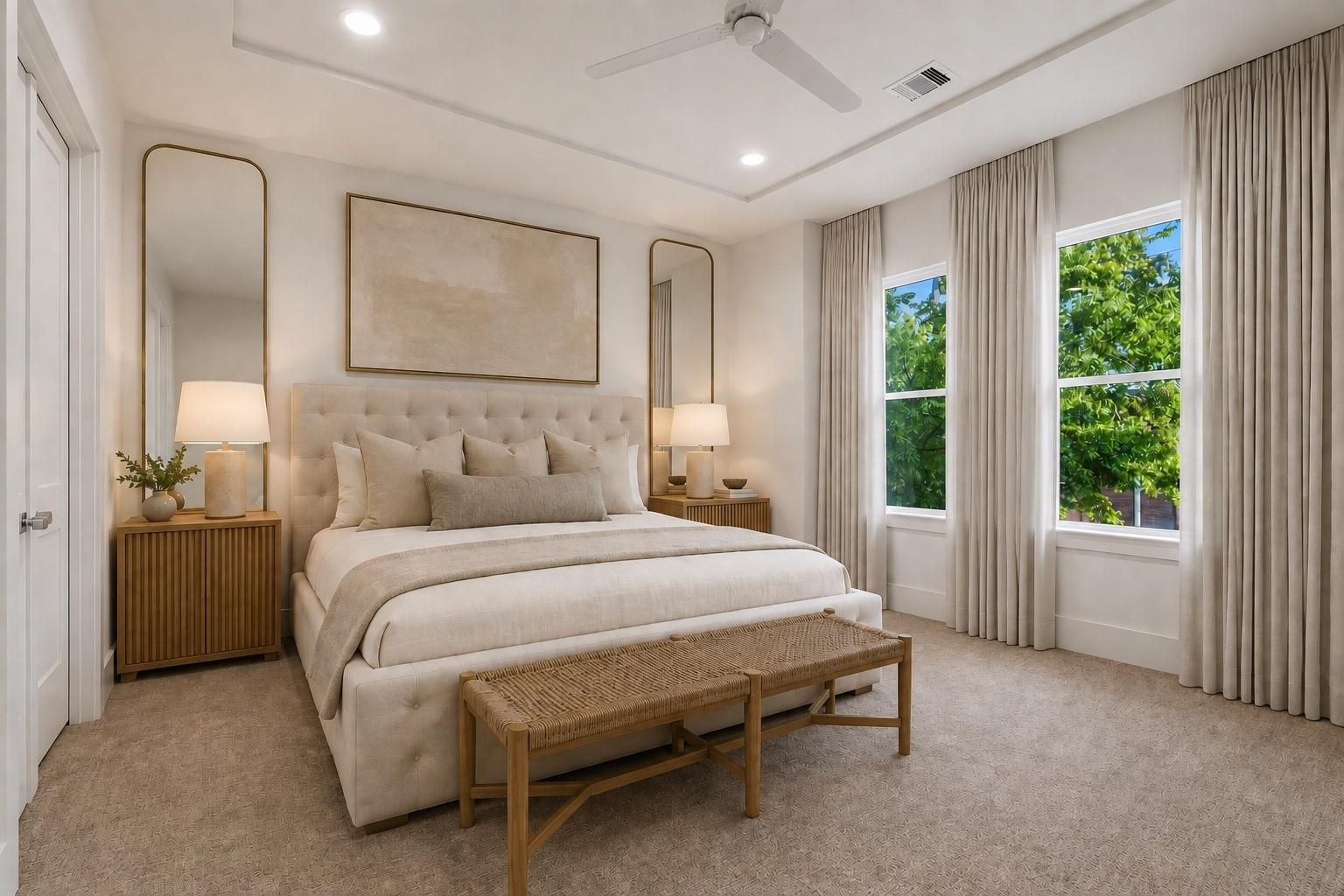 Staged bedroom with an upholstered bed, woven bench, tall mirrors, and leafy views through two windows.