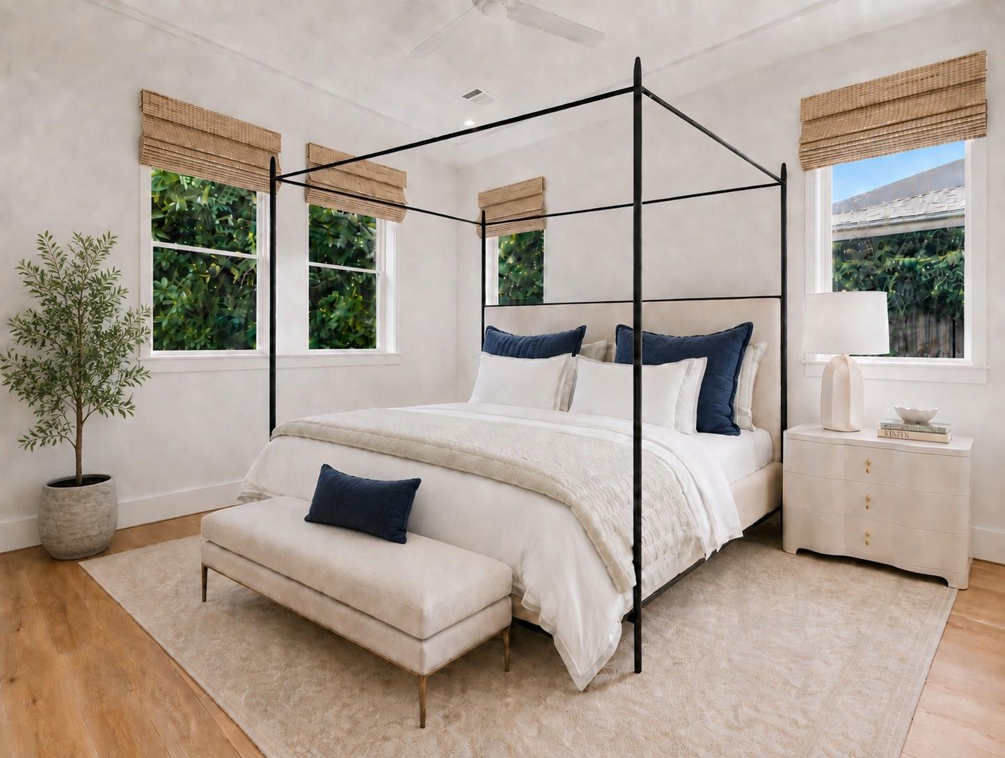 Bedroom with a black canopy bed, navy pillows, and a cream bench beneath woven-shade windows.
