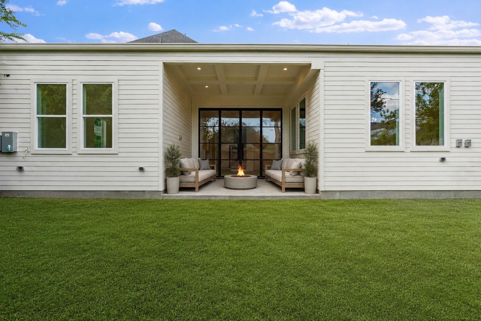 White-sided house with a covered patio, two sofas, a fire pit, and a broad green lawn.