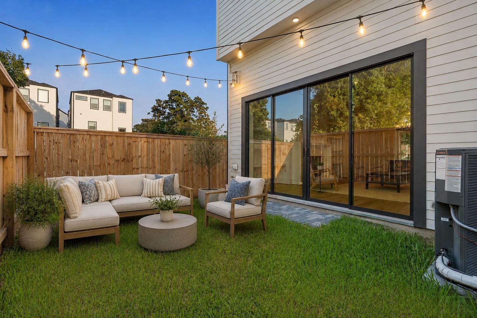 Backyard patio with cushioned lounge seating, sliding glass doors, and glowing string lights.