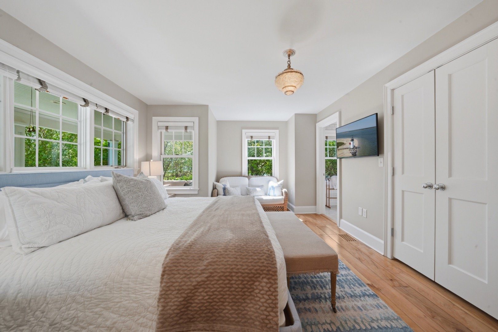 Furnished bedroom with a white quilted bed, multiple windows, and a small sitting area.