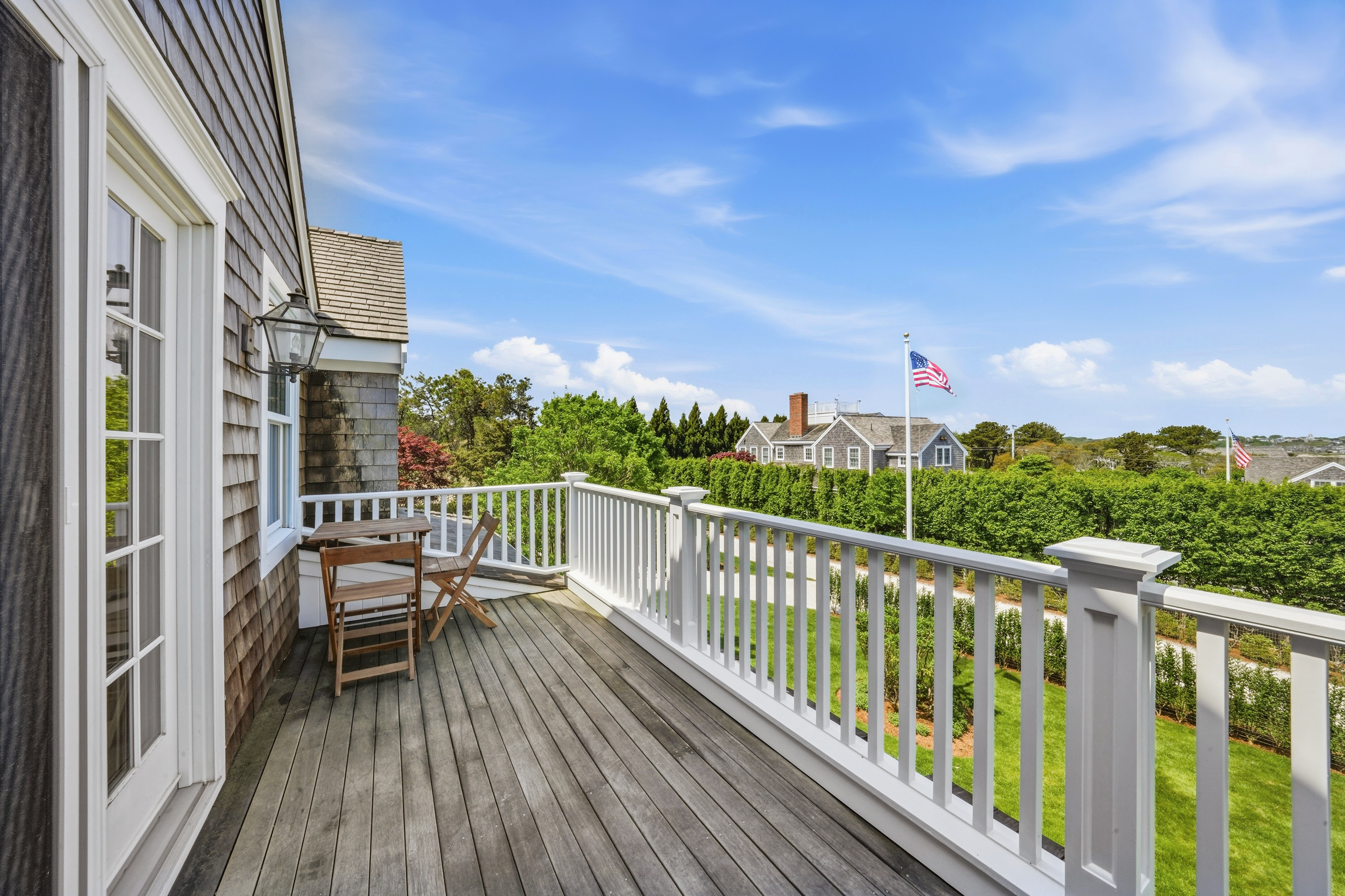 Wood deck with white railings and chairs overlooking green hedges and neighboring homes.