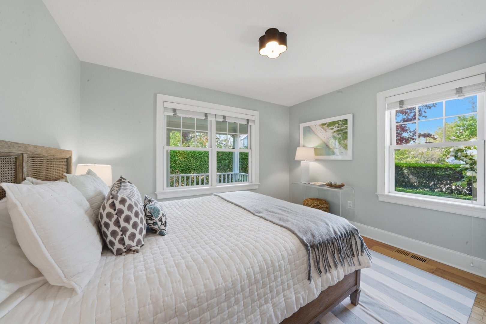 Bedroom with a white quilted bed and two windows overlooking green hedges.