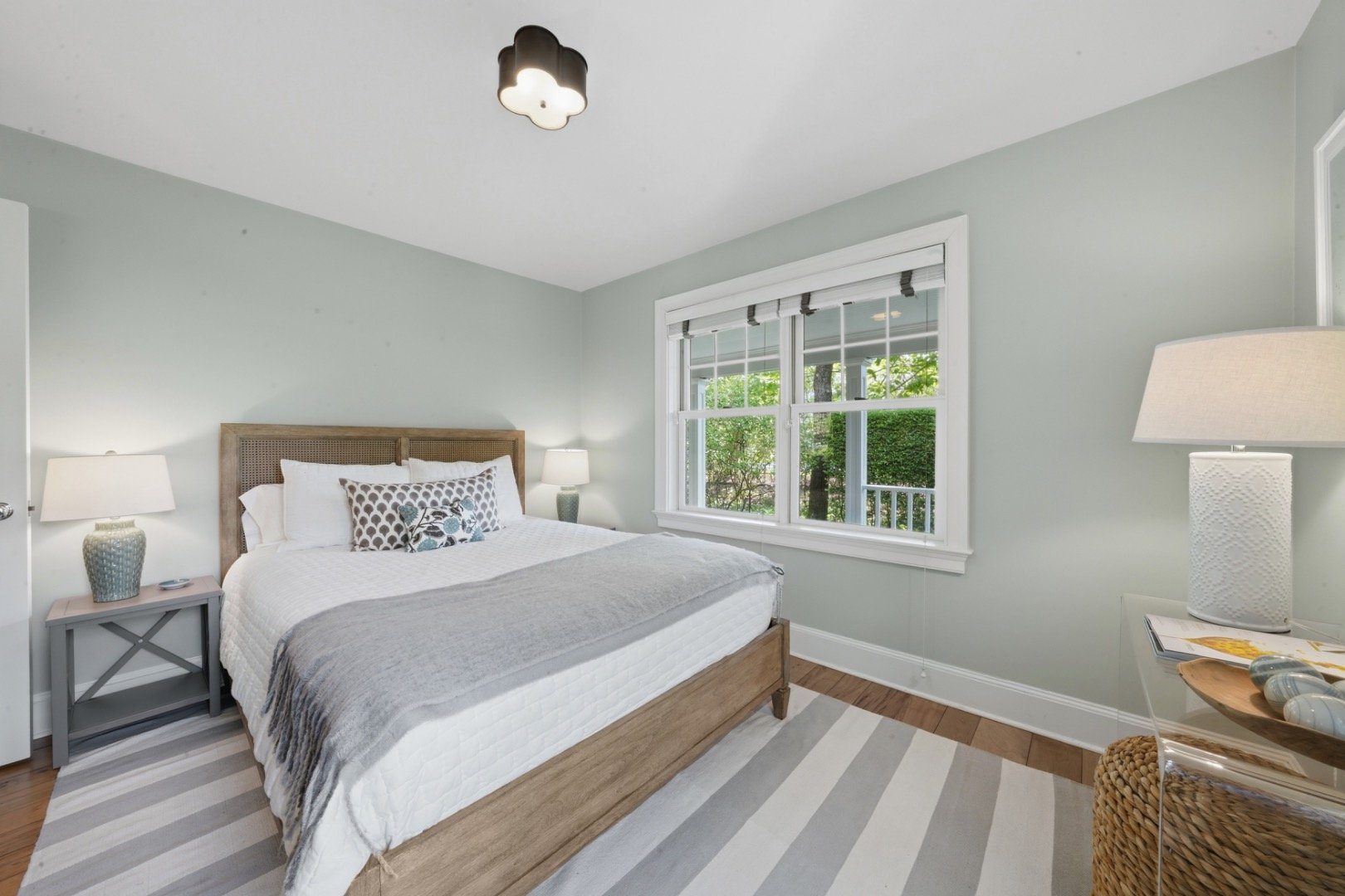 Bedroom with a woven-headboard bed, pale green walls, and a large window overlooking greenery.