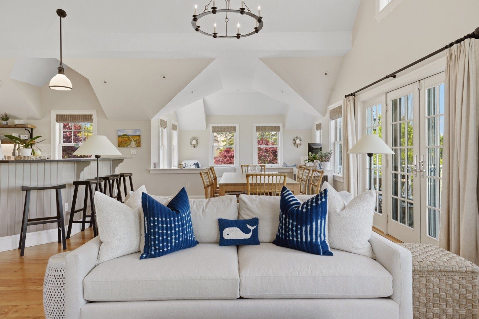 Open-plan living and dining room with a white sofa, blue pillows, vaulted ceiling, and French doors.