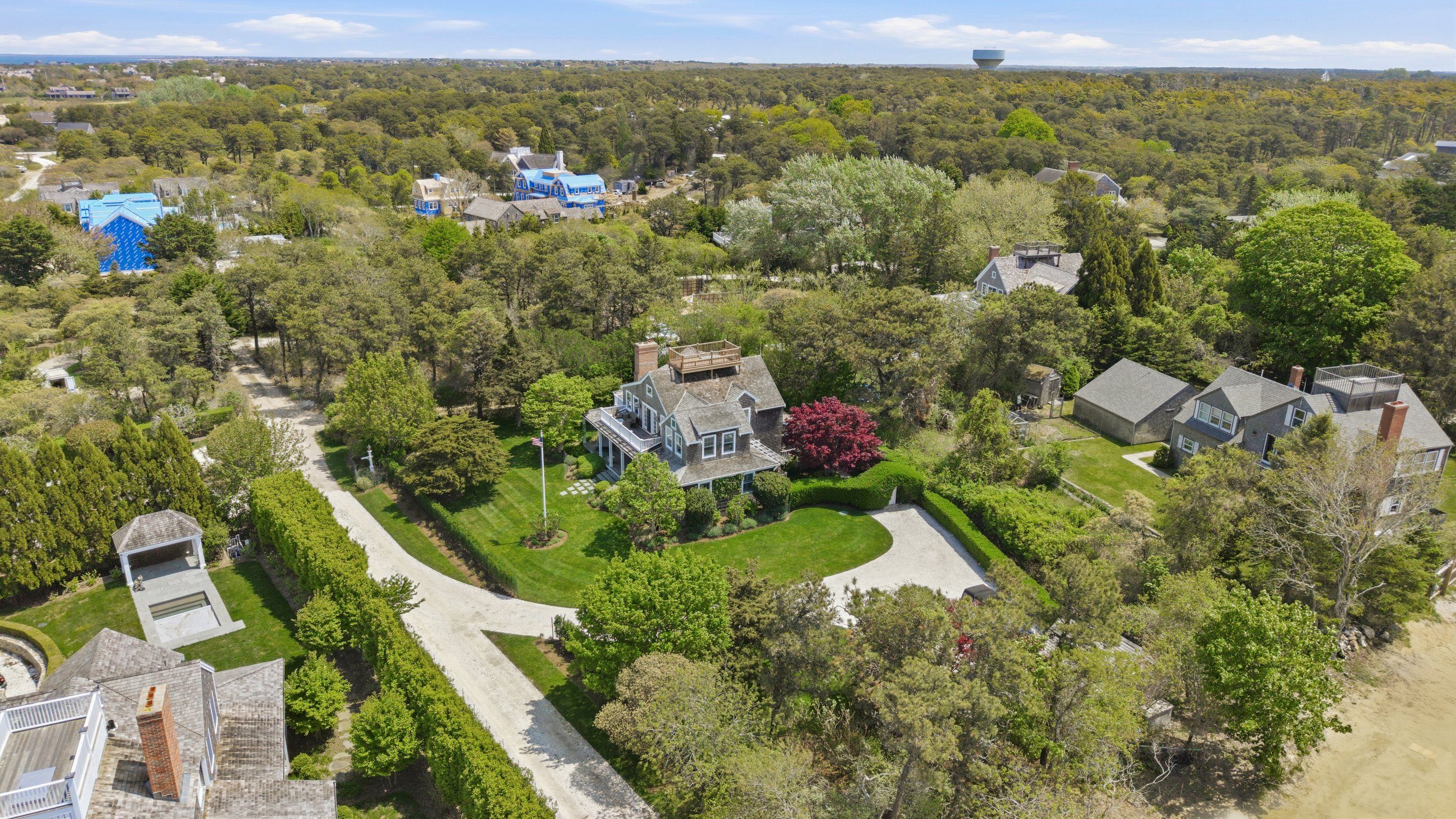 Aerial view of a shingled home with a rooftop deck, curved driveway, and leafy grounds near the shore.