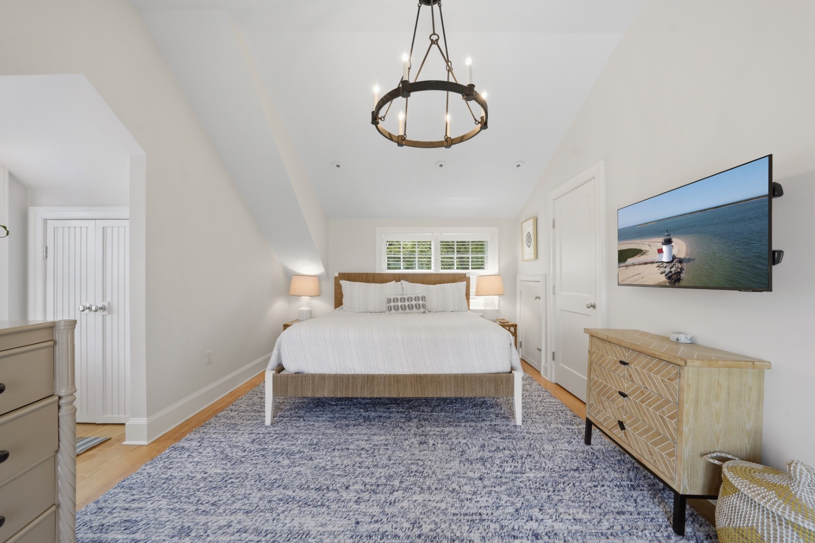 Furnished bedroom with a white bed, blue patterned rug, and television displaying a lighthouse.