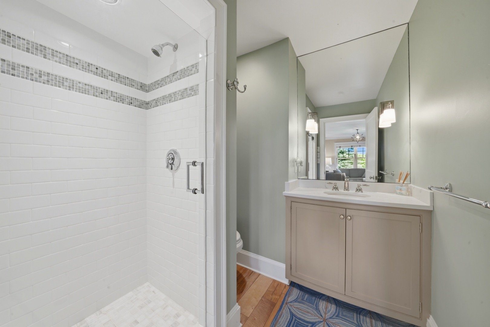 Bathroom with white tiled shower, gray-accent bands, taupe vanity, and blue patterned rug.