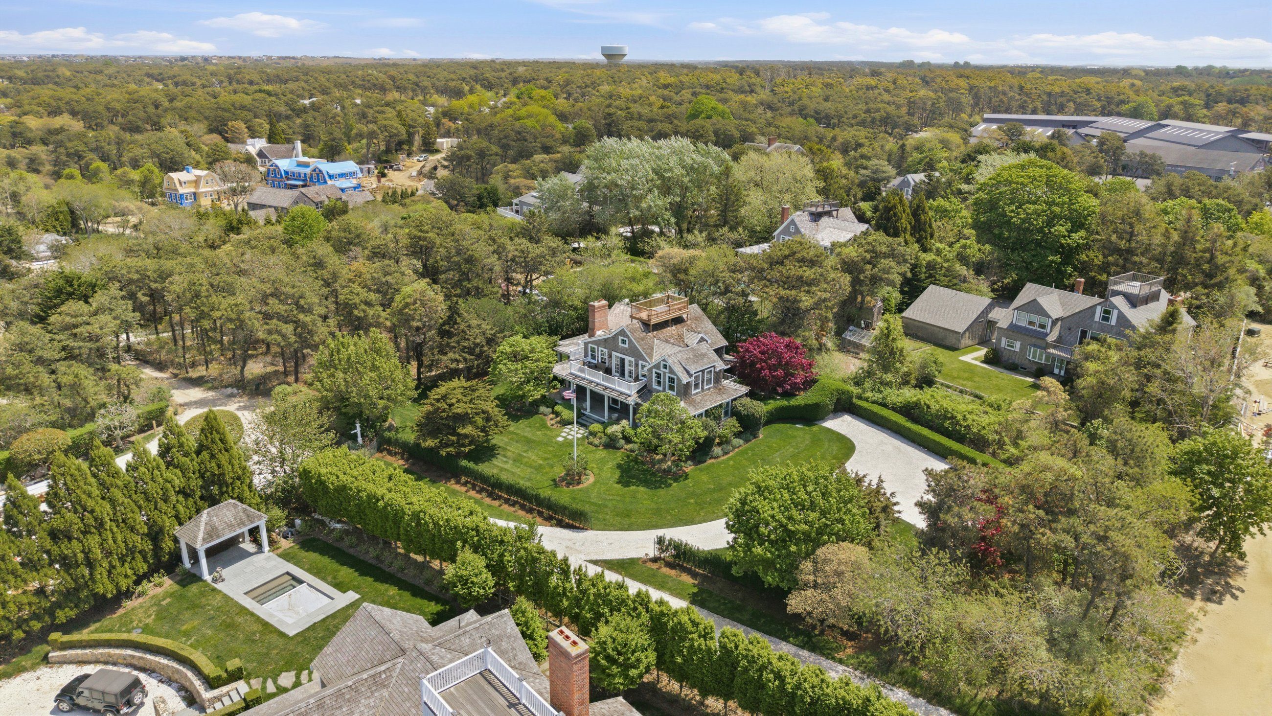 Aerial view of a gray-shingled home among trees, with landscaped grounds, a curved driveway, and nearby sandy shoreline.