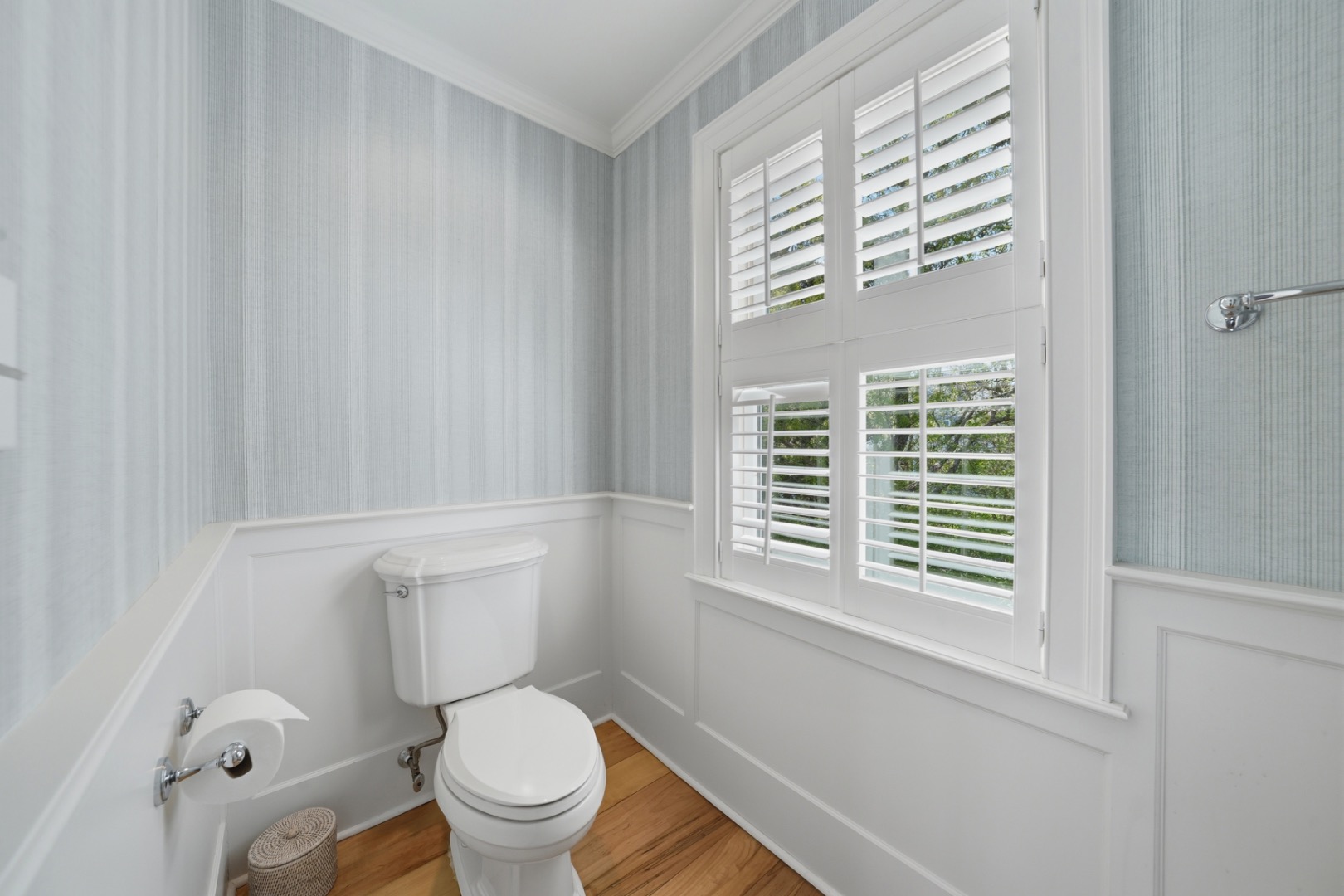 Bathroom with a white toilet, plantation-shuttered window, pale striped walls, and warm wood flooring.