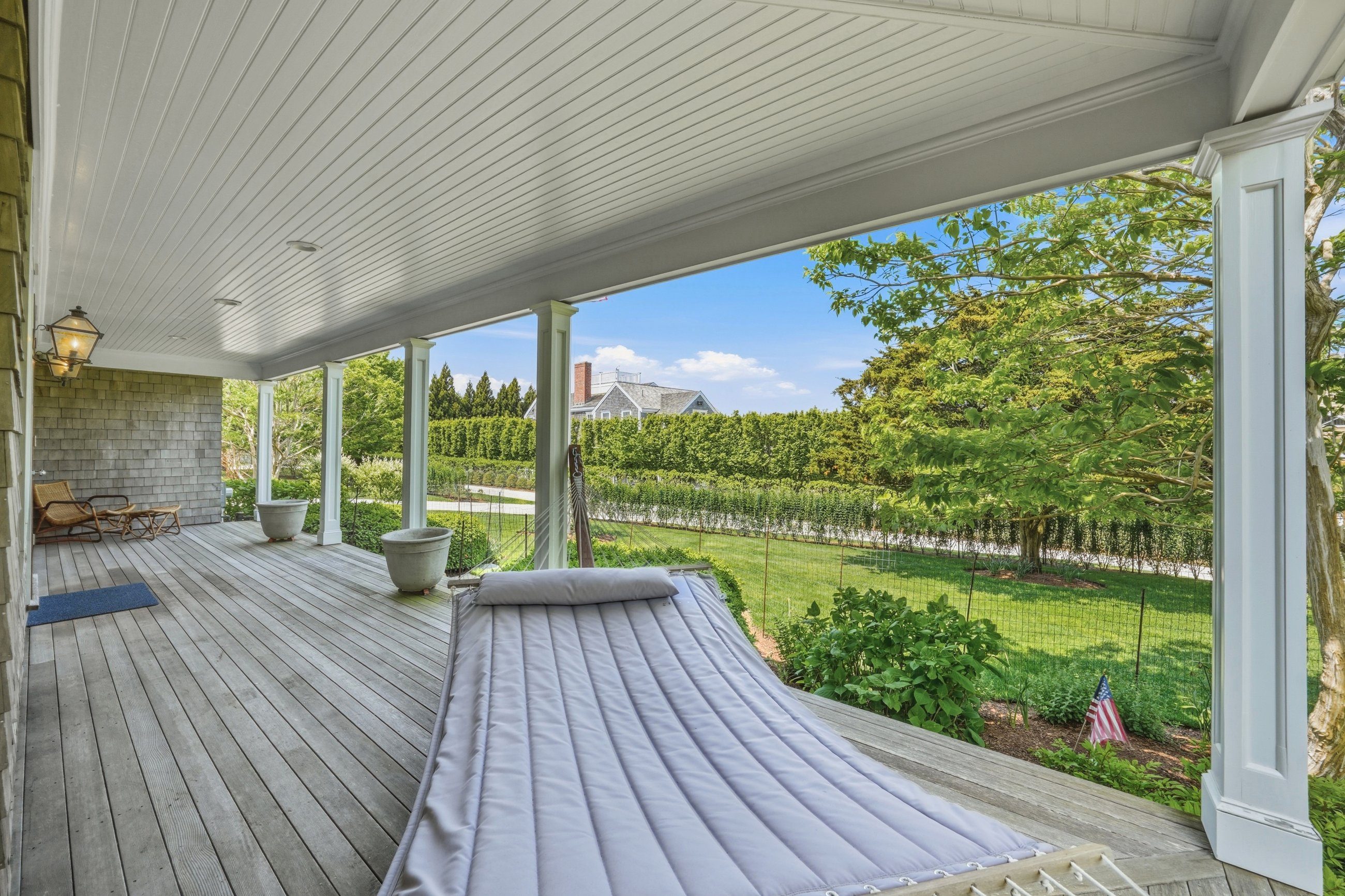 Covered porch with a cushioned swing overlooking a lawn bordered by hedges and trees.