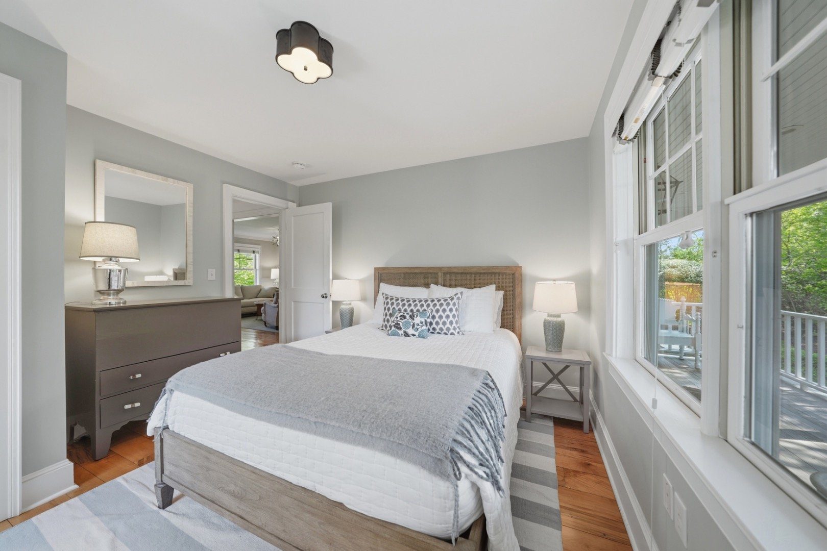 Bedroom with a white bed, gray throw, and tall windows overlooking a deck and greenery.