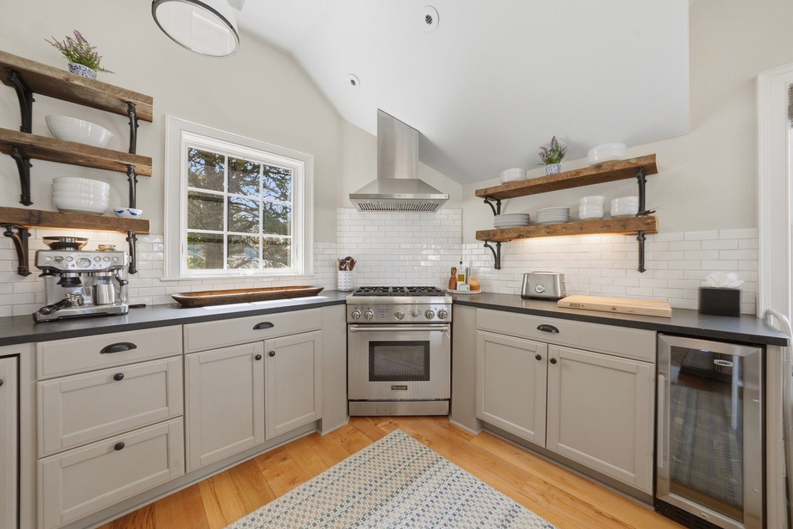 Kitchen with vaulted ceiling, rustic open shelves, stainless-steel range, and window overlooking trees.