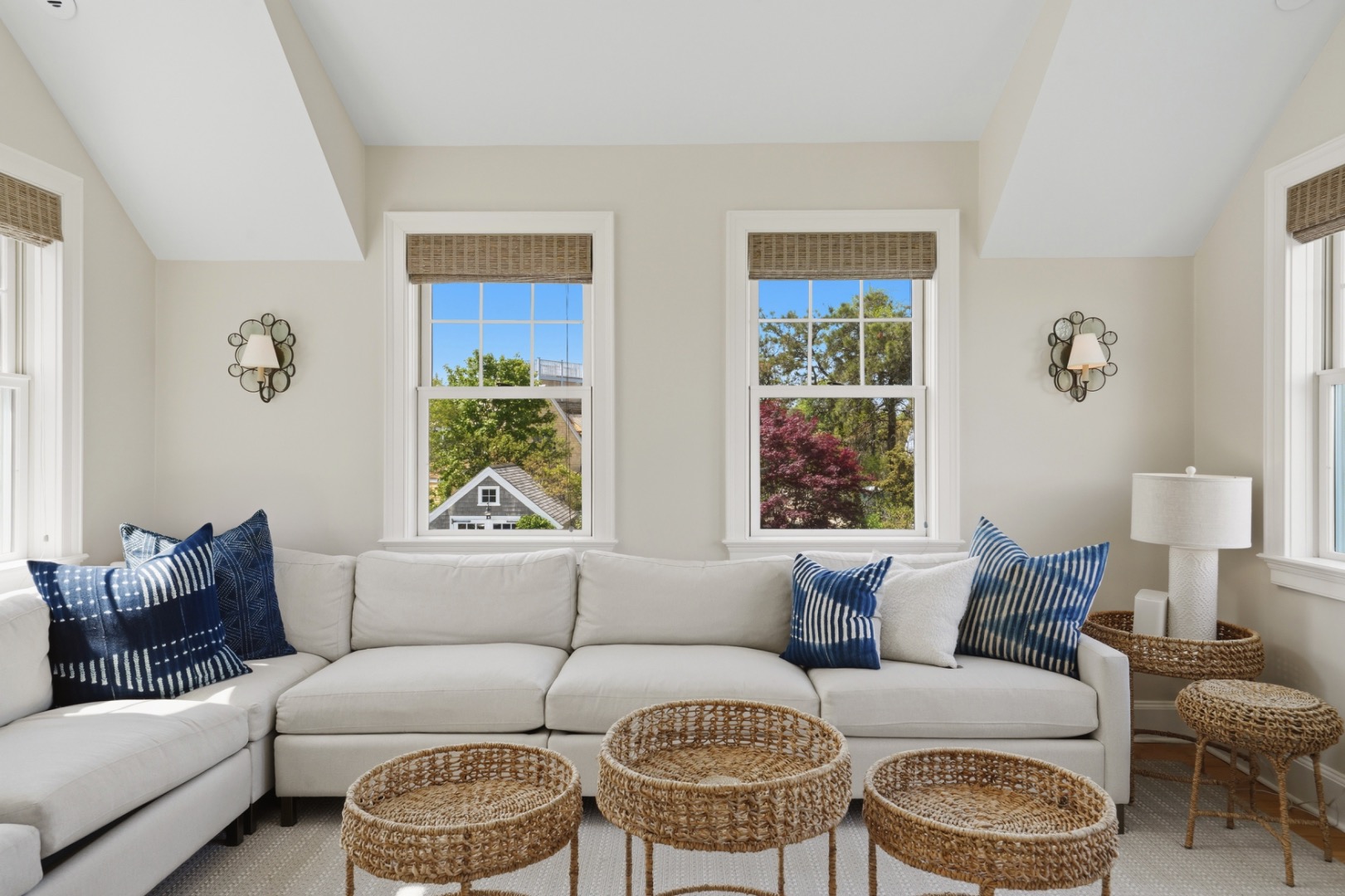 Living room with a white sectional, navy pillows, woven tables, and large windows overlooking trees.
