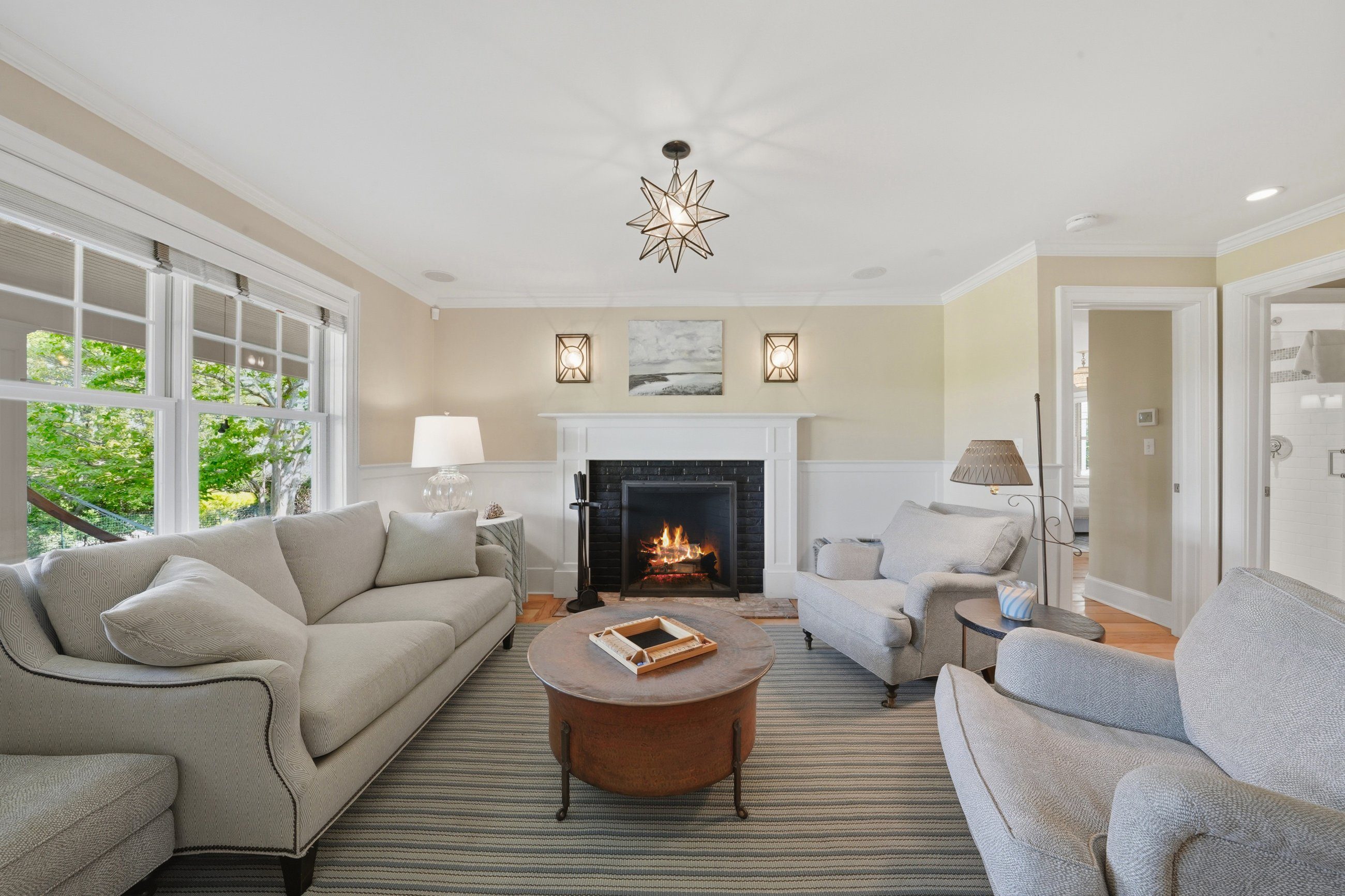 Living room with a lit fireplace, pale sofas, and large windows overlooking leafy trees.
