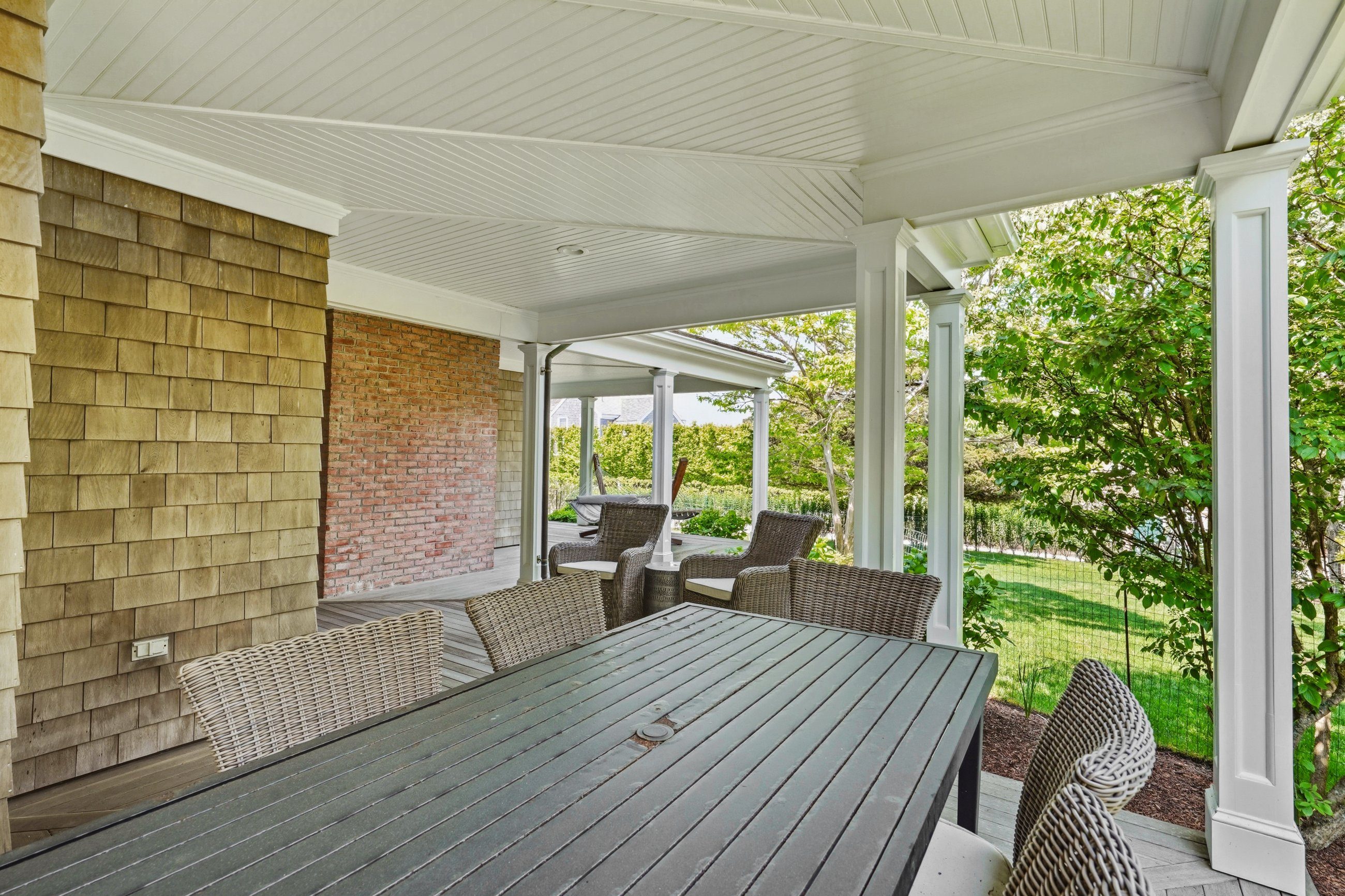 Covered porch with wicker seating and a large dining table beside a leafy garden.